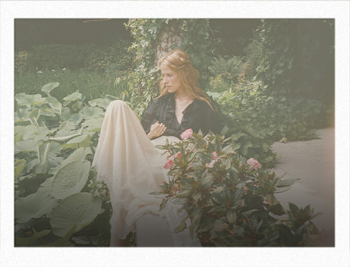 A woman with blonde hair sits outdoors in a garden setting, wearing a black top and a flowing cream-colored skirt. Large green leaves and pink flowers frame the scene, with a soft, vintage-toned filter over the photograph.