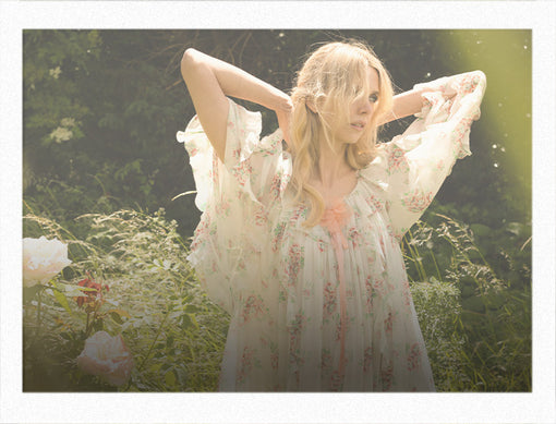 Woman in a floral dress standing in a natural setting with sunlight filtering through.