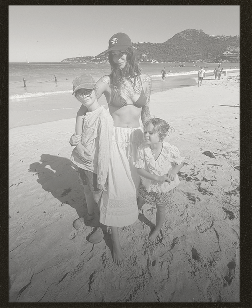 Black and white photo of a woman and two children on a beach with mountains in the background.