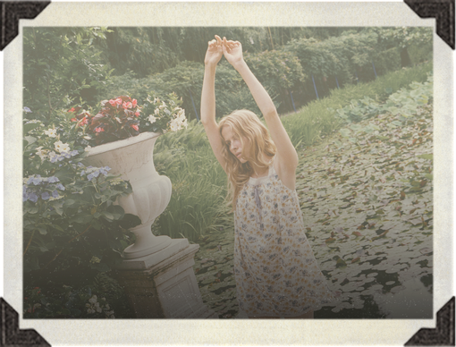 A woman in a light floral print dress stands in a garden with her arms raised above her head, next to a large white urn planter.