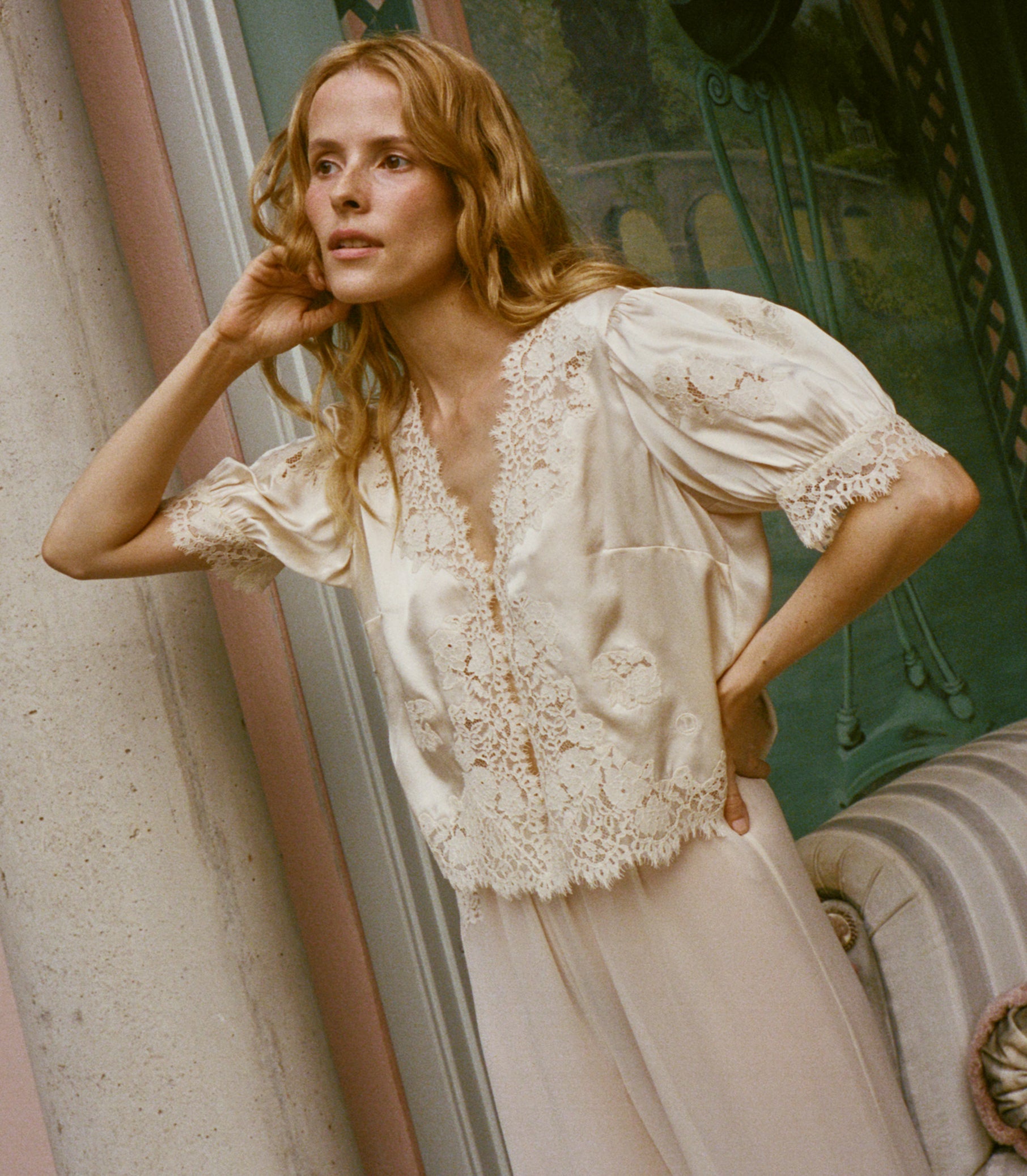 A woman wearing a cream-colored lace-trimmed silk blouse with puff sleeves and cream pants, posed in front of a vintage-style window. 1
