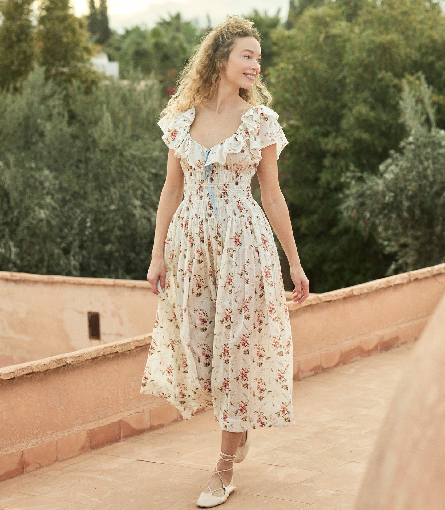 Woman in a white floral dress standing on a deck with greenery in the background