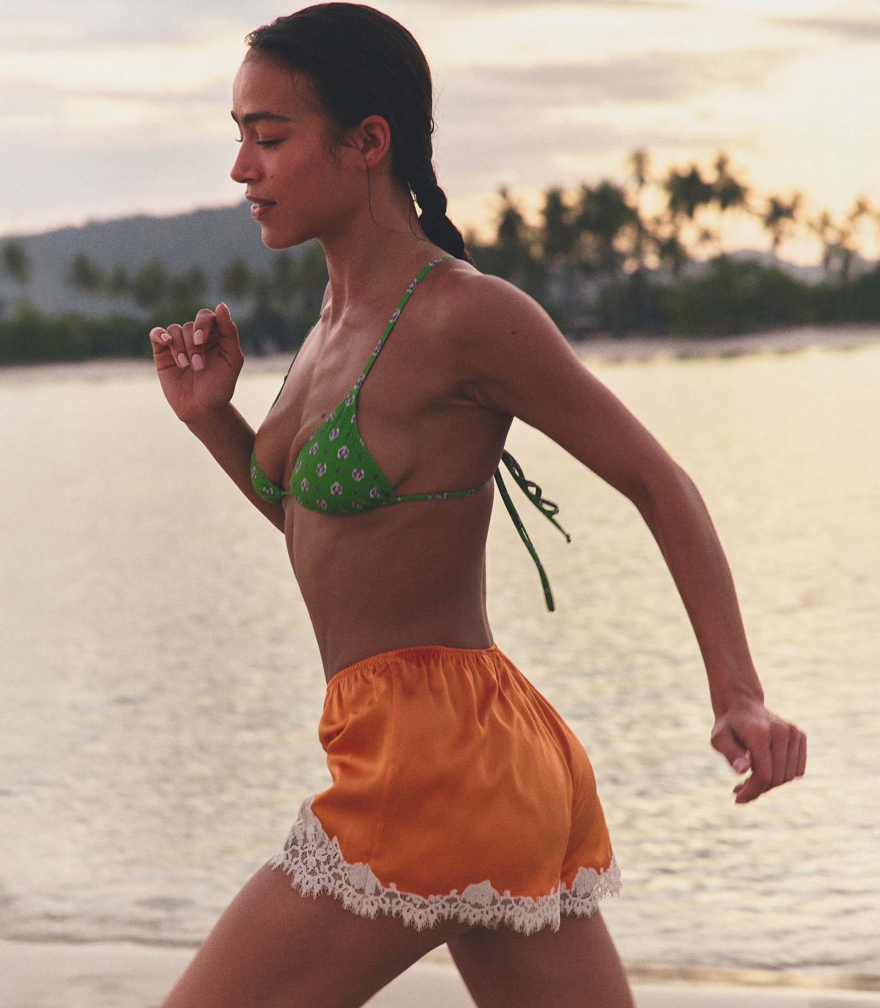 Woman in green bikini top and orange shorts standing on a beach at sunset. 1
