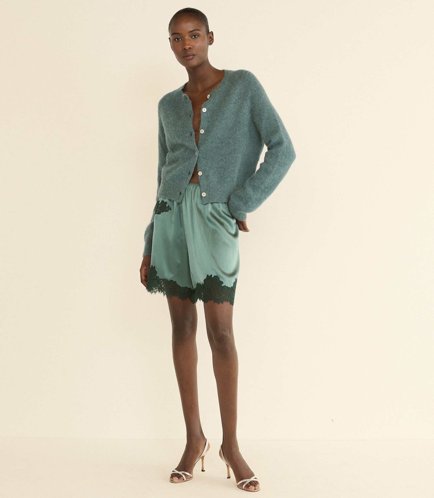 A model stands against a plain cream studio background wearing a sage green cardigan, a matching sage green satin pair of shorts with dark green lace trim, and beige high-heeled sandals. 1
