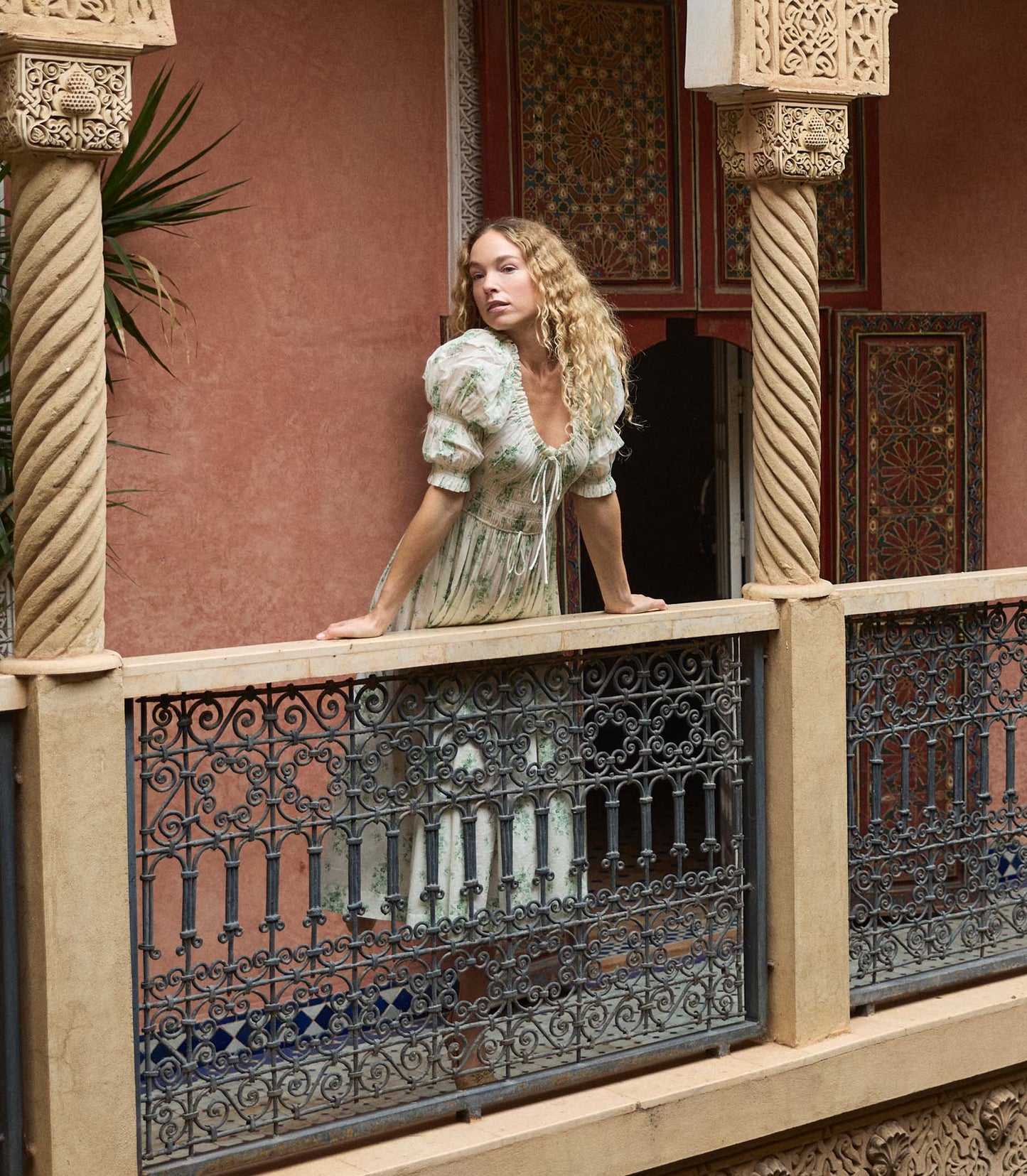 Woman standing on a balcony with ornate columns and railing, wearing a light-colored floral dress.