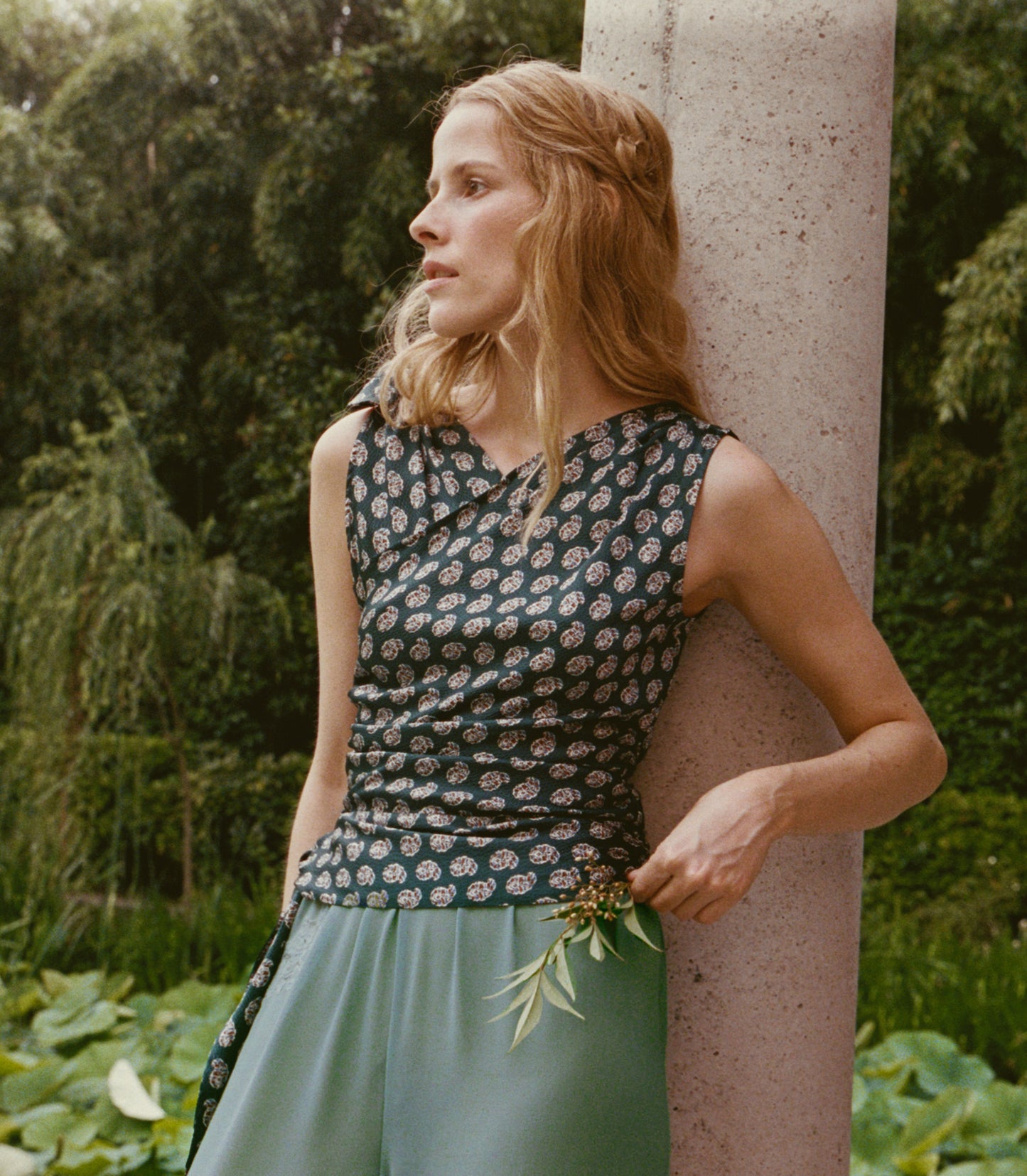 A woman in a sleeveless dark paisley top and sage green pants leans against a stone pillar in a lush garden setting.