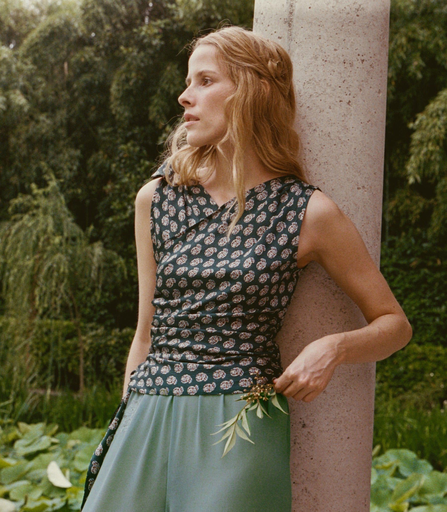 A woman in a sleeveless dark paisley top and sage green pants leans against a stone pillar in a lush garden setting.