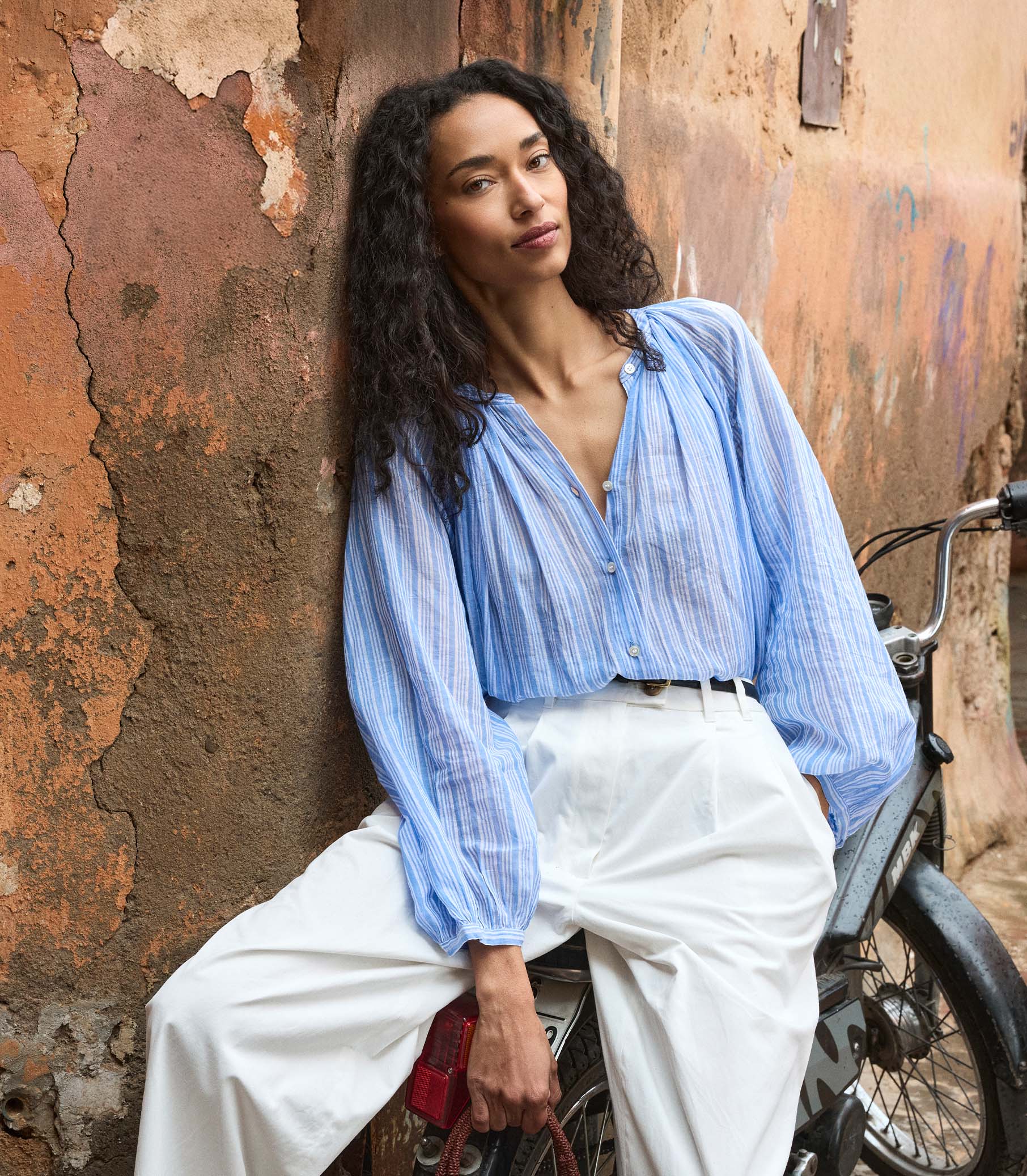 Woman in a striped blue shirt and white pants sitting on a motorcycle against a textured wall. 6
