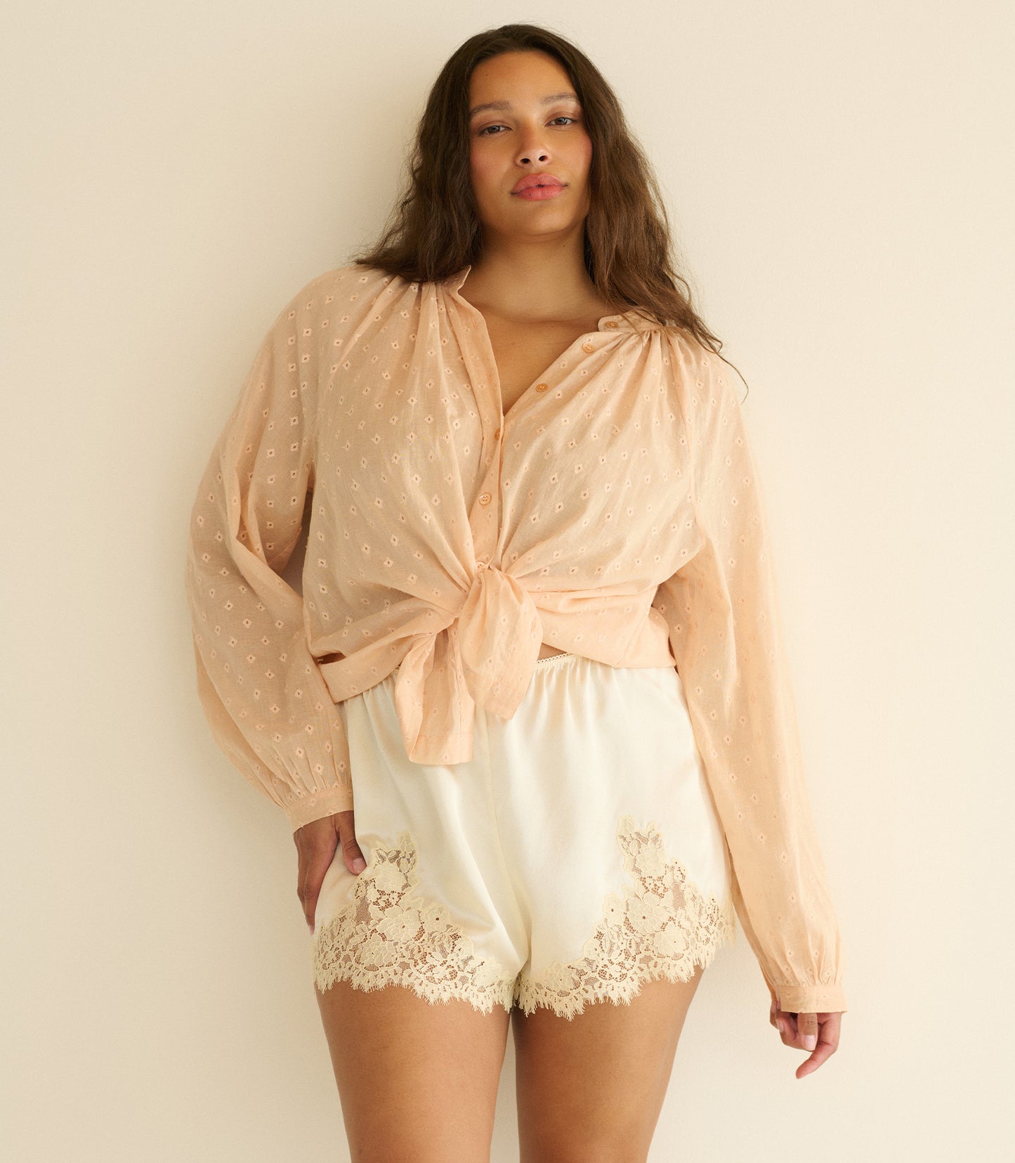 Woman wearing a light pink top and white lace shorts against a beige background