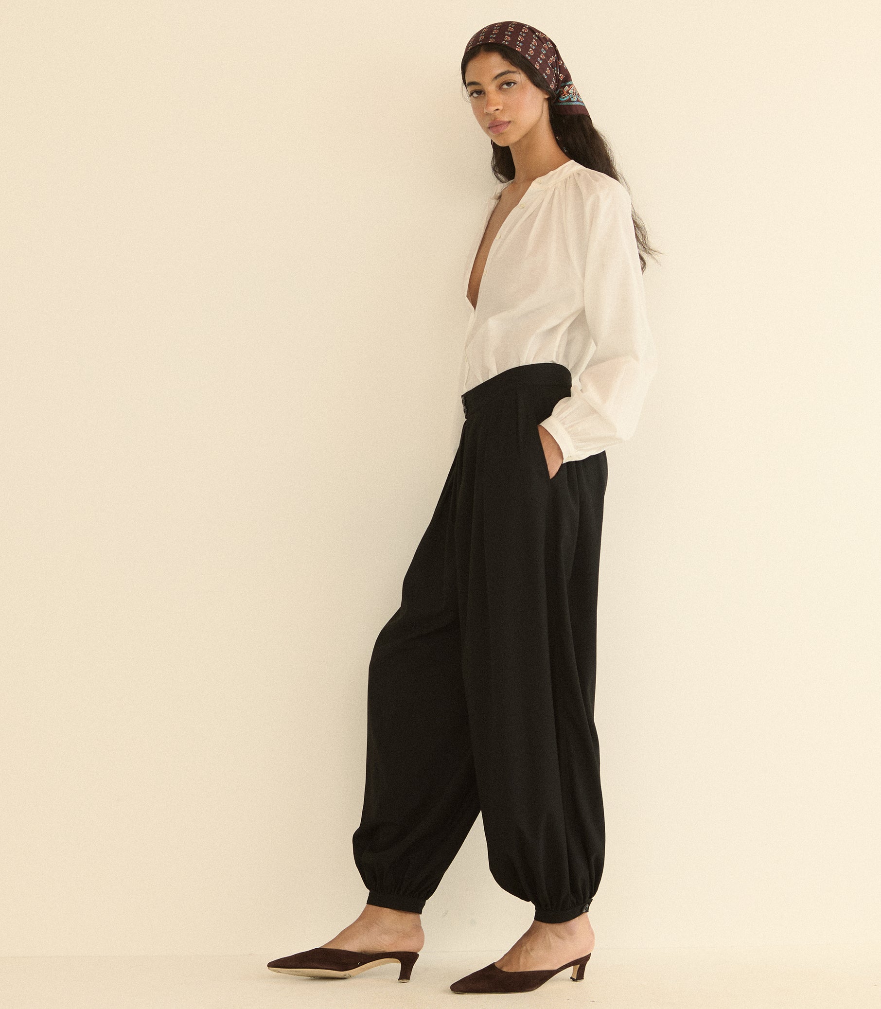 A woman modeling loose black harem-style pants with a white blouse and brown pointed-toe mule heels, standing against a plain warm off-white studio background.