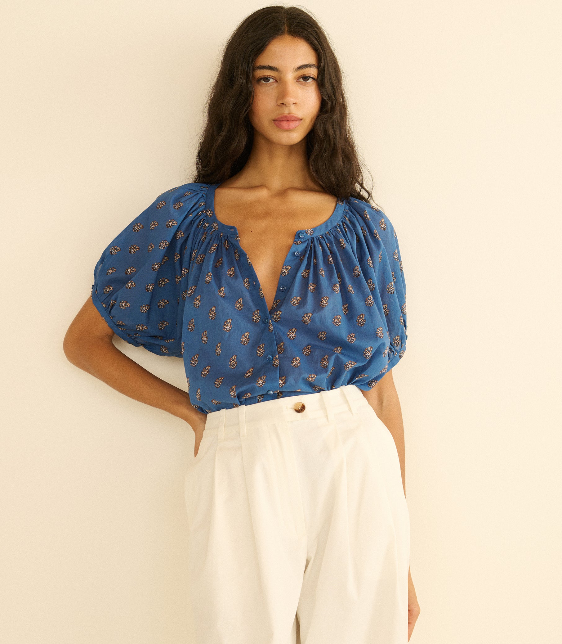 Woman wearing a blue paisley blouse with white pants against a beige background