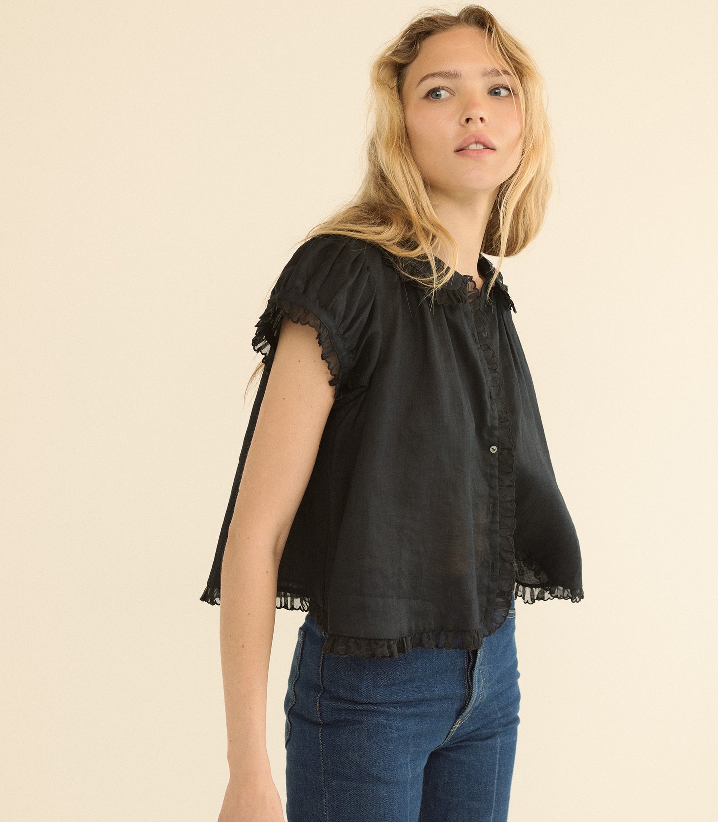 A woman wearing a black short-sleeve blouse with ruffled trim and blue jeans, photographed against a plain beige studio background.