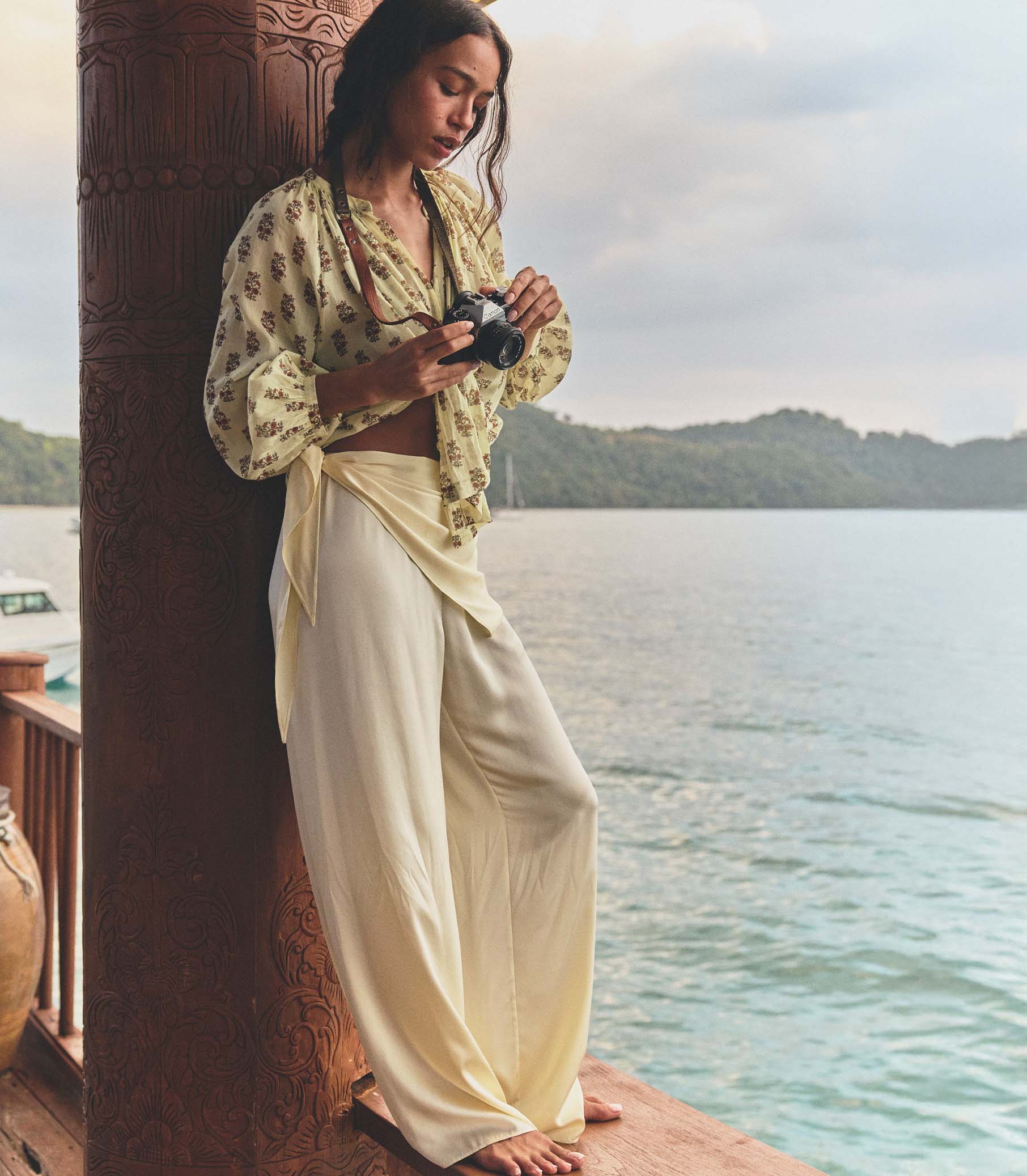 Woman in a yellow top and white pants holding a camera by a waterfront. 1
