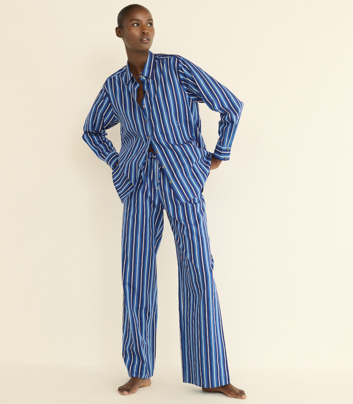 A model stands barefoot against a plain cream background wearing a matching two-piece pajama set with vertical blue and white stripes. Sporting a long-sleeve button-front top with a split neckline and wide-leg pants.