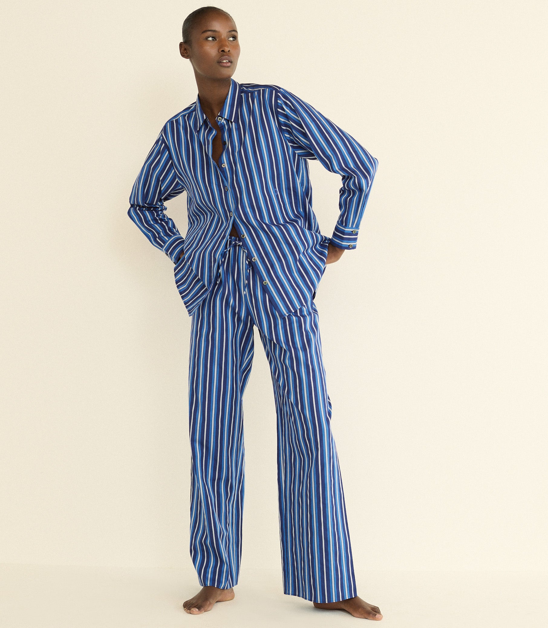 A model stands barefoot against a plain cream background wearing a matching two-piece pajama set with vertical blue and white stripes. Sporting a long-sleeve button-front top with a split neckline and wide-leg pants. 1
