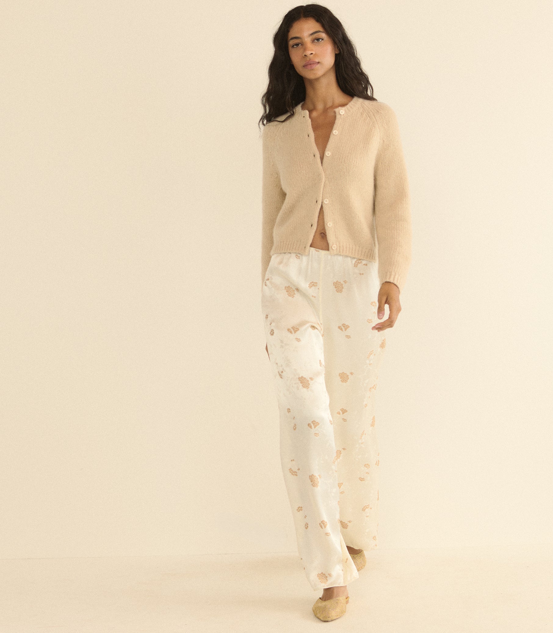 A woman modeling a cozy two-piece loungewear set consisting of a beige button-up cardigan and white pants with a light tan floral pattern, set against a solid cream background.