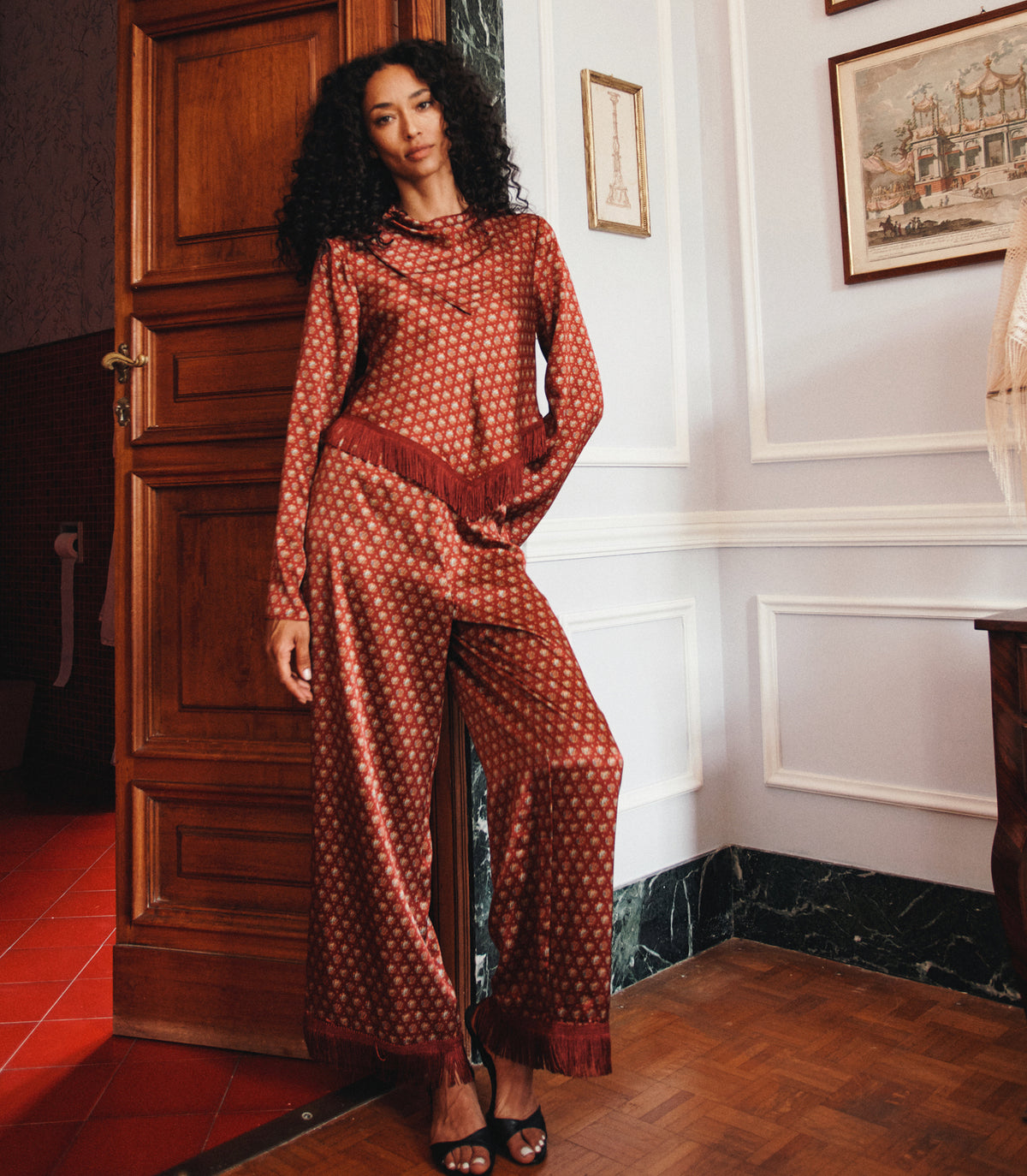 A woman models a rust-orange and white patterned two-piece set consisting of a long-sleeve top and matching wide-leg pants with fringe details, standing in an elegant room with wooden paneling and a dark wood door. 1
