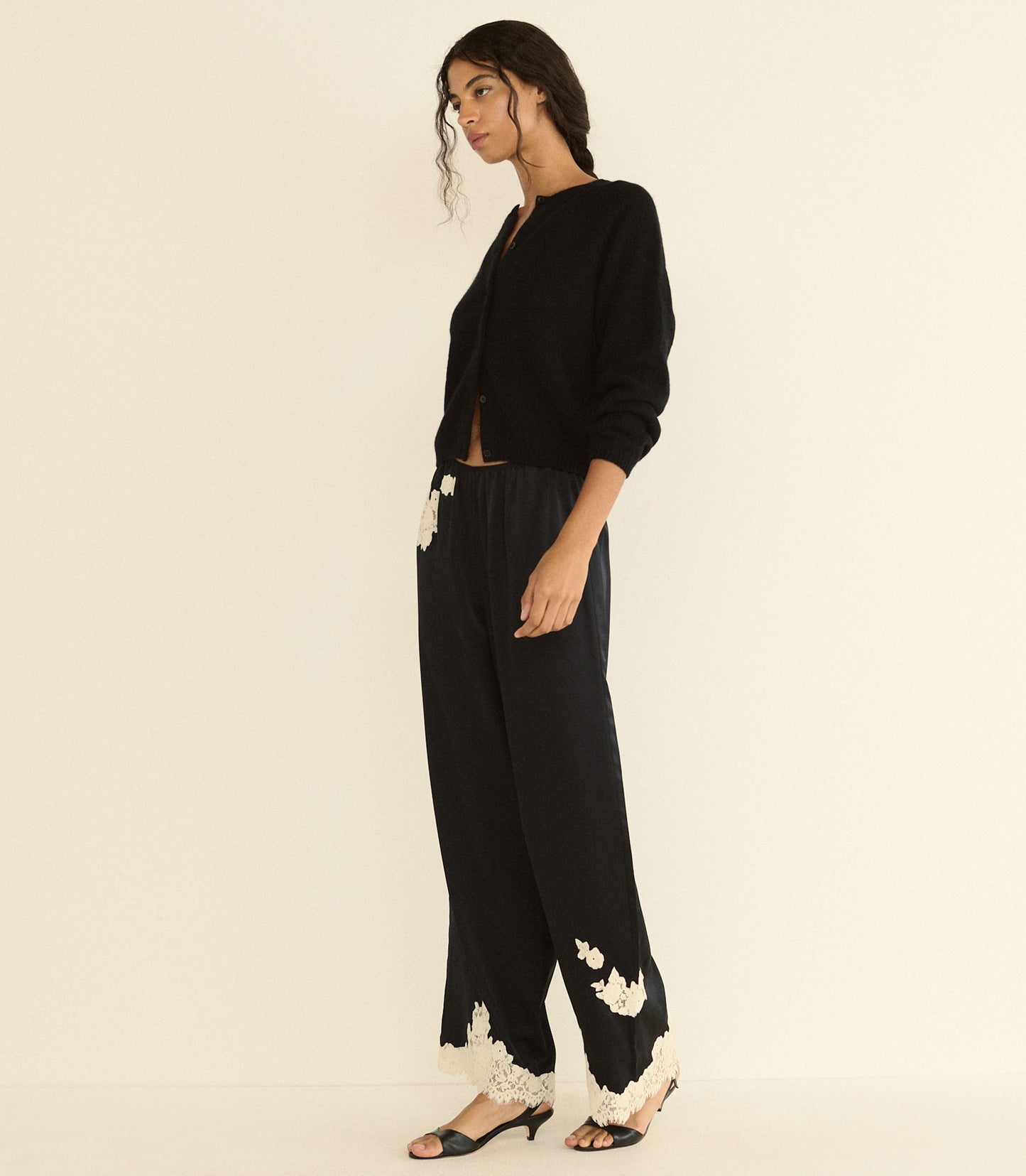 A woman models a black cardigan and wide-leg black trousers with white floral lace trim at the hems, paired with black heeled sandals, against a plain cream background.