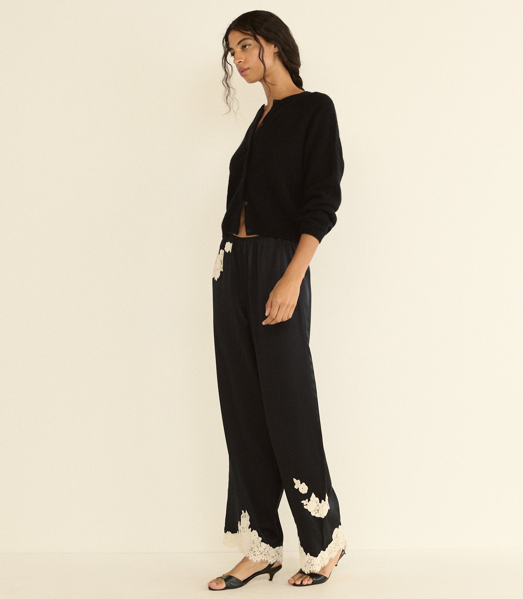 A woman models a black cardigan and wide-leg black trousers with white floral lace trim at the hems, paired with black heeled sandals, against a plain cream background. 1
