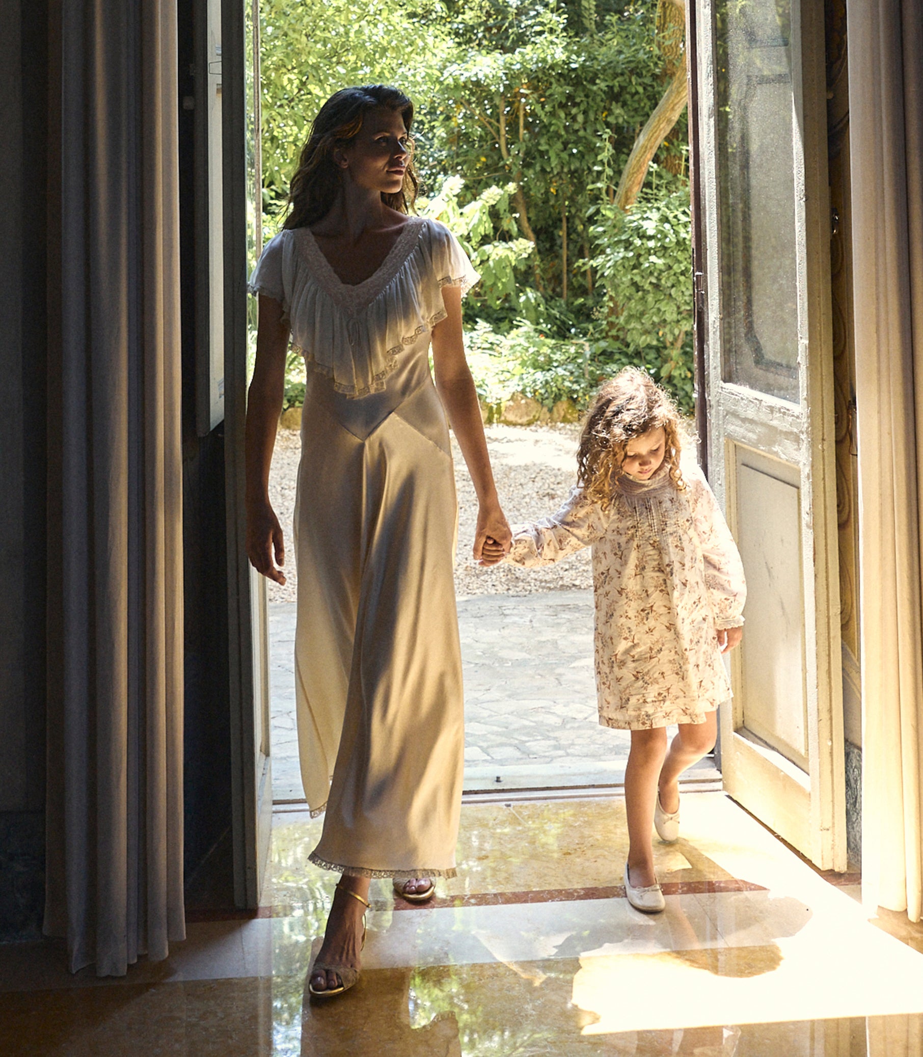 A woman and a young girl walk hand in hand out of a sunlit room through open doors, wearing matching light-colored dresses. The woman wears a cream satin slip dress with fringe at the neckline, and the girl wears a matching cream floral print dress. 1
