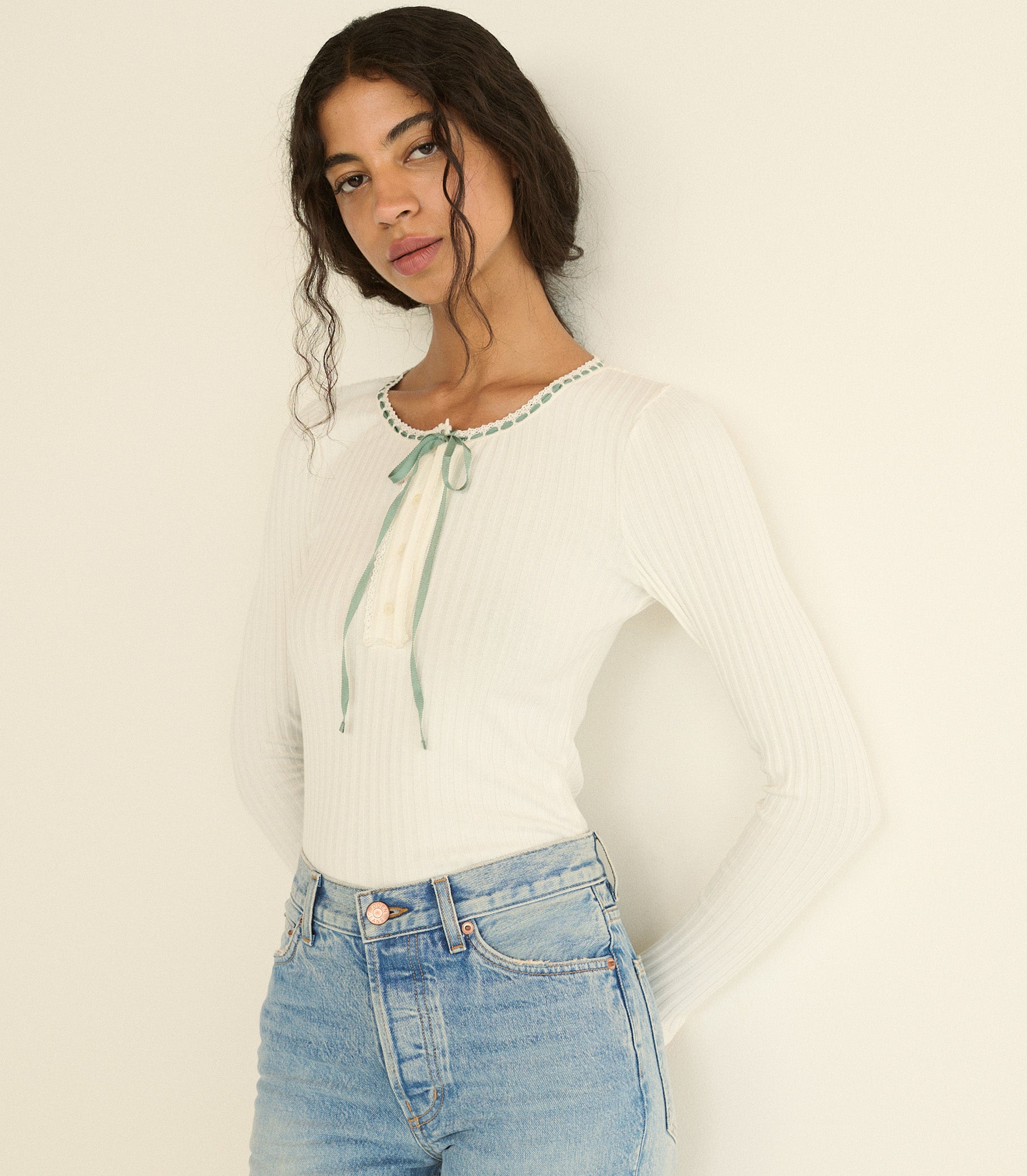 A woman wearing a white ribbed long-sleeve top with a mint blue tie detail at the neckline, paired with light blue jeans, against a plain off-white background.