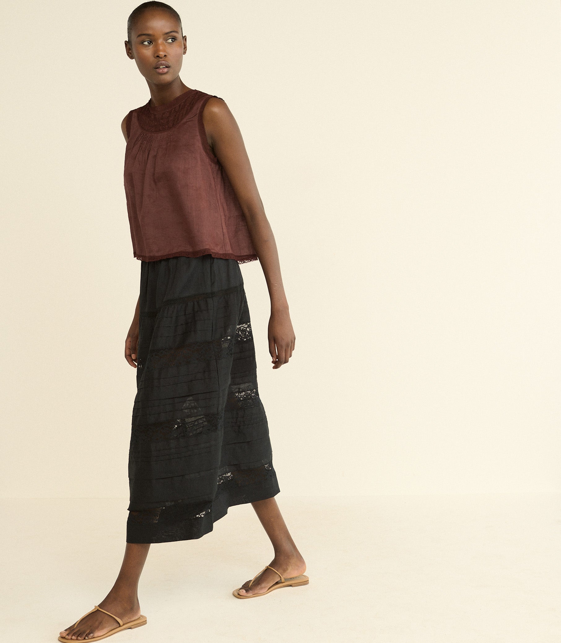 A model wearing a sleeveless rust-brown top and a black tiered midi skirt with distressed details, paired with flat sandals, against a solid cream background.
