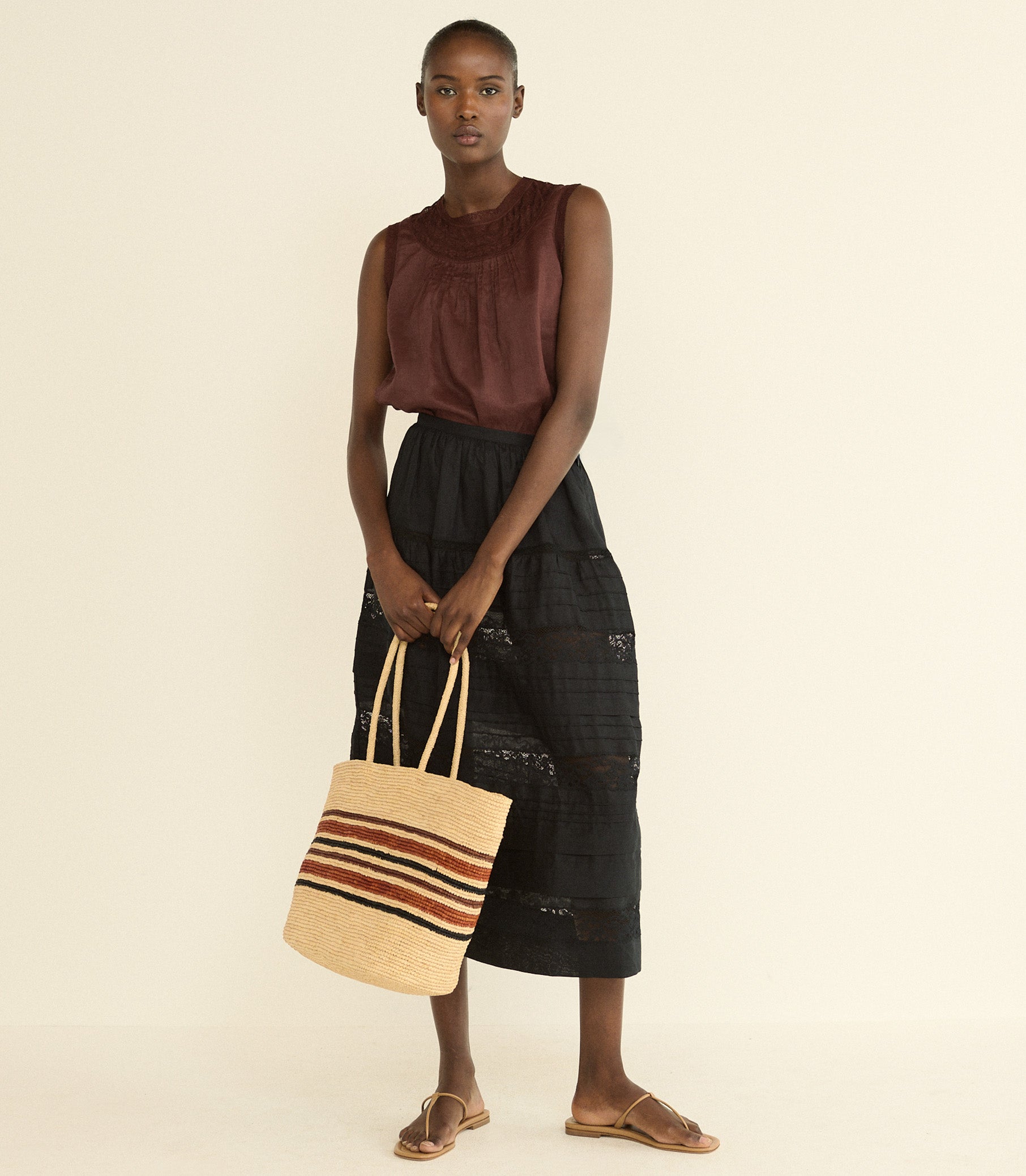 A model stands against a plain cream background wearing a sleeveless rust-brown top, a black patterned midi skirt, and brown sandals, while holding a large woven straw tote bag with orange, brown, and cream stripes.