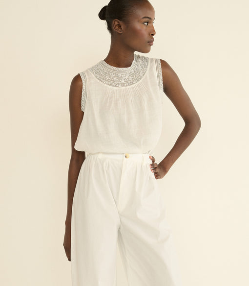 A model wearing a white sleeveless top with a detailed embroidered yoke and matching white wide-leg trousers, photographed against a solid light beige background.