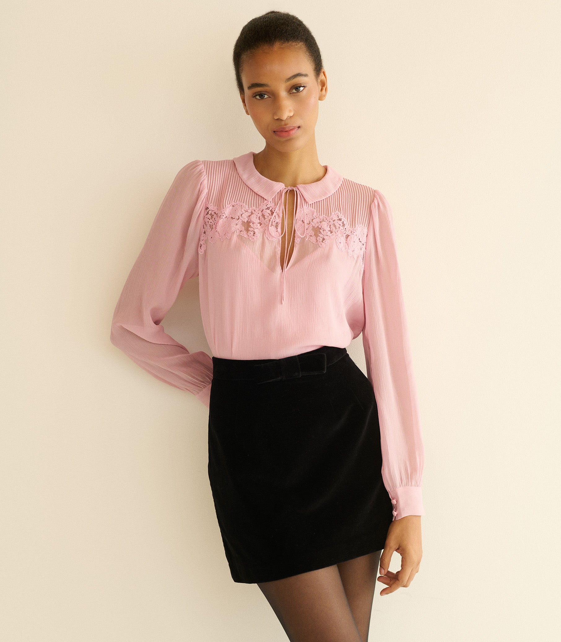 Woman wearing a pink blouse with floral details and a black skirt against a beige background 1
