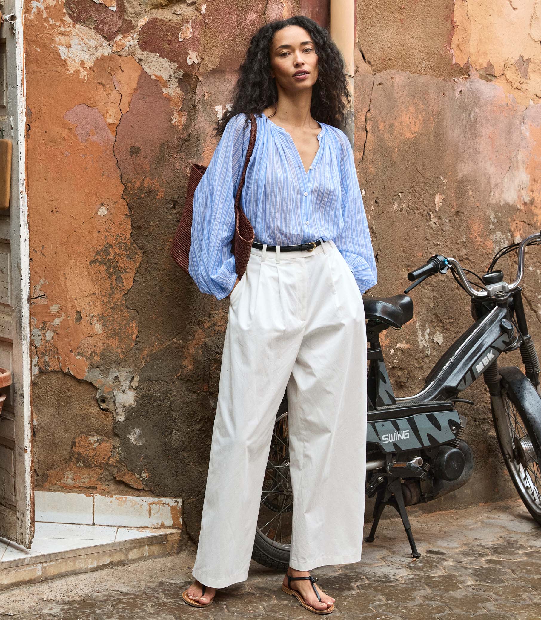 Woman in a striped blue shirt and white pants standing next to a motorcycle against a textured wall. 4
