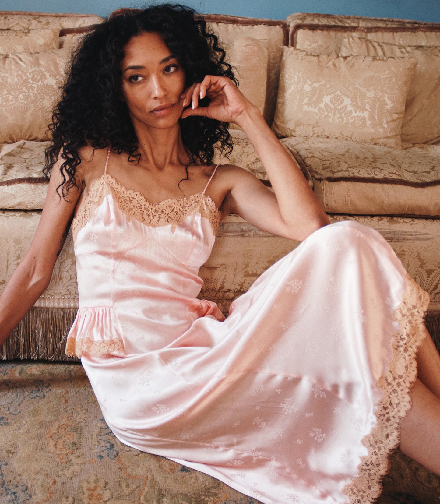 A woman with curly black hair reclines on a beige sofa wearing a light pink satin slip dress with lace trim and a subtle floral pattern.