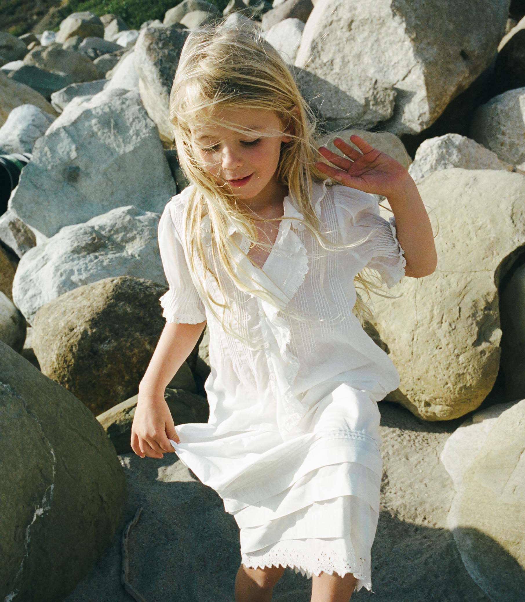 Young girl in a white blouse and white skirt sitting among large rocks. 1
