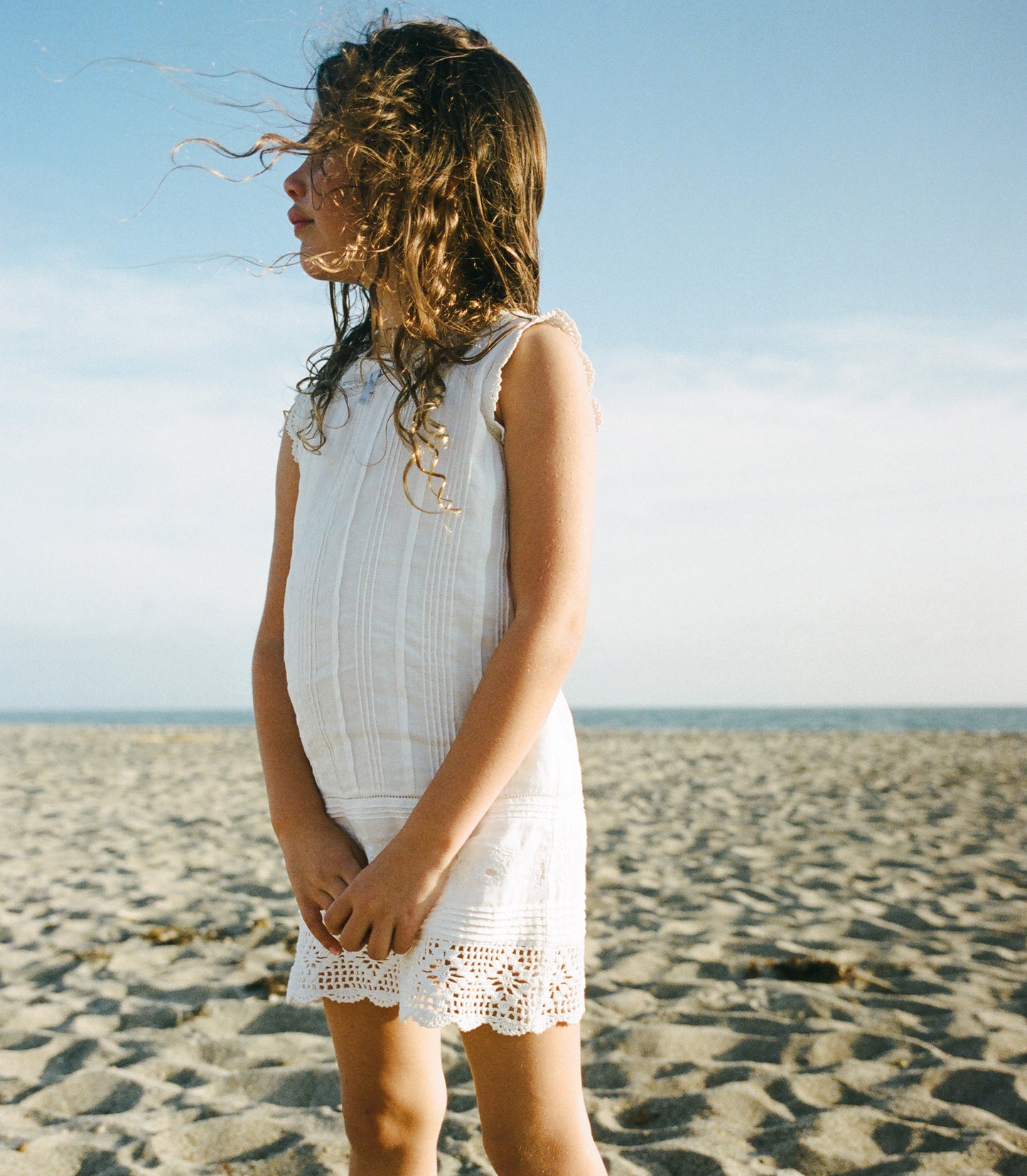 Child in a white dress standing on a sandy beach with a clear sky.