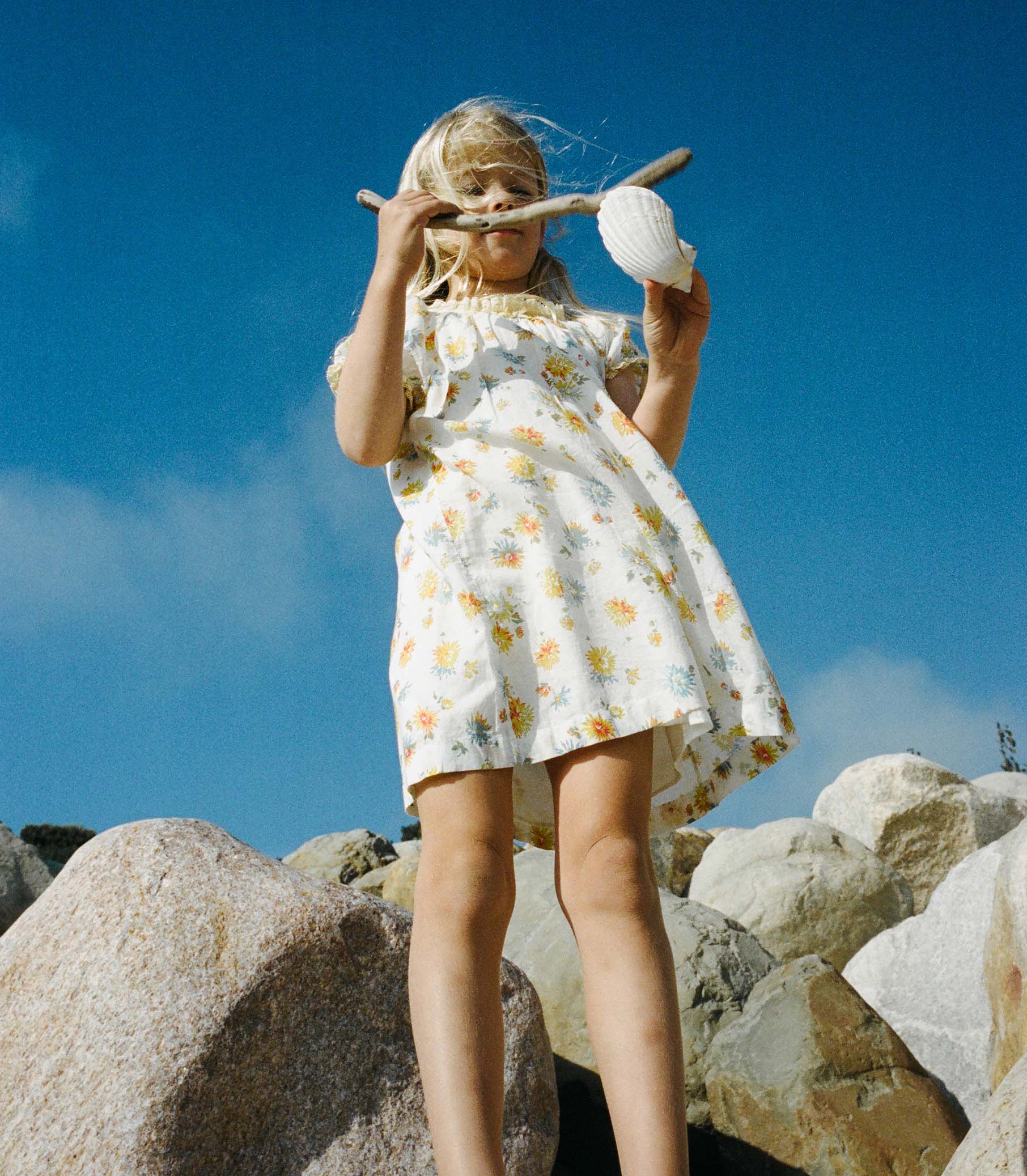 Young girl in a white floral dress holding a shell and a stick on rocky terrain with a blue sky. 1
