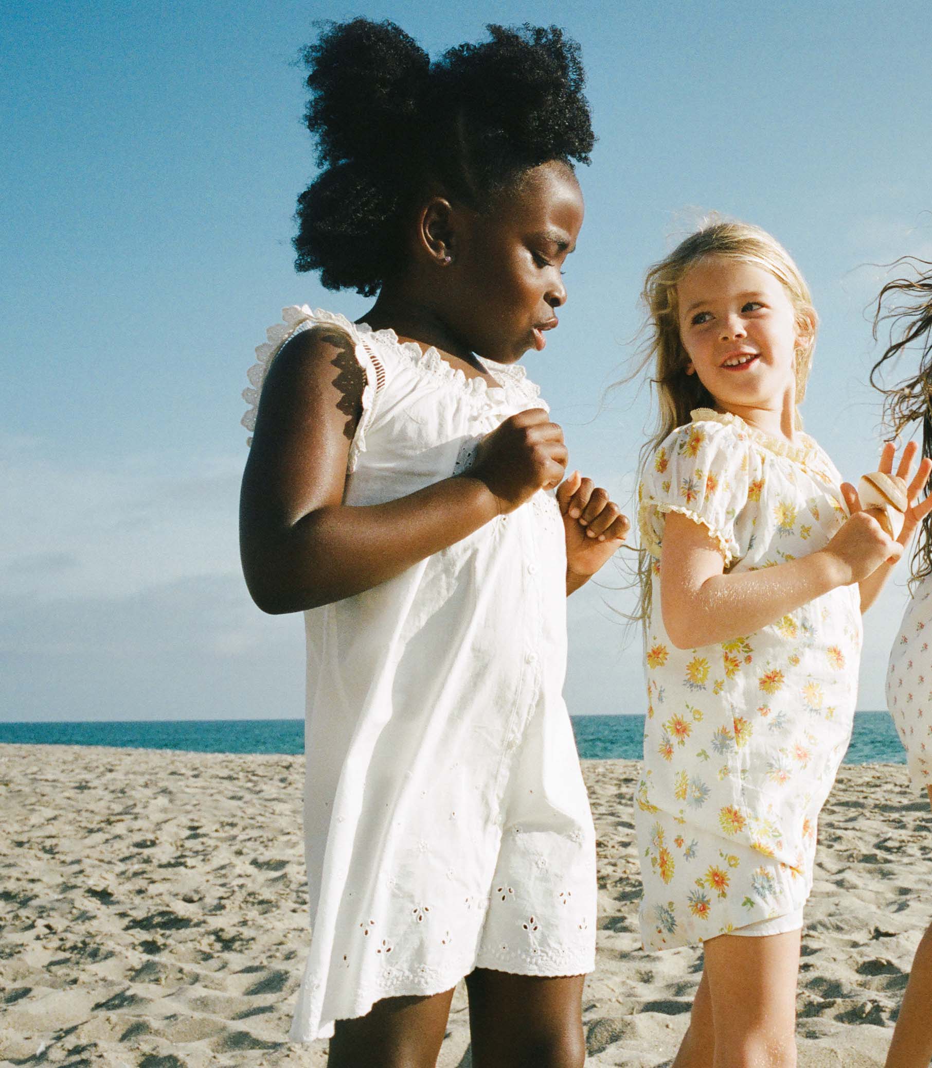 Two young girls in white dresses standing on a sandy beach with blue sky. 1
