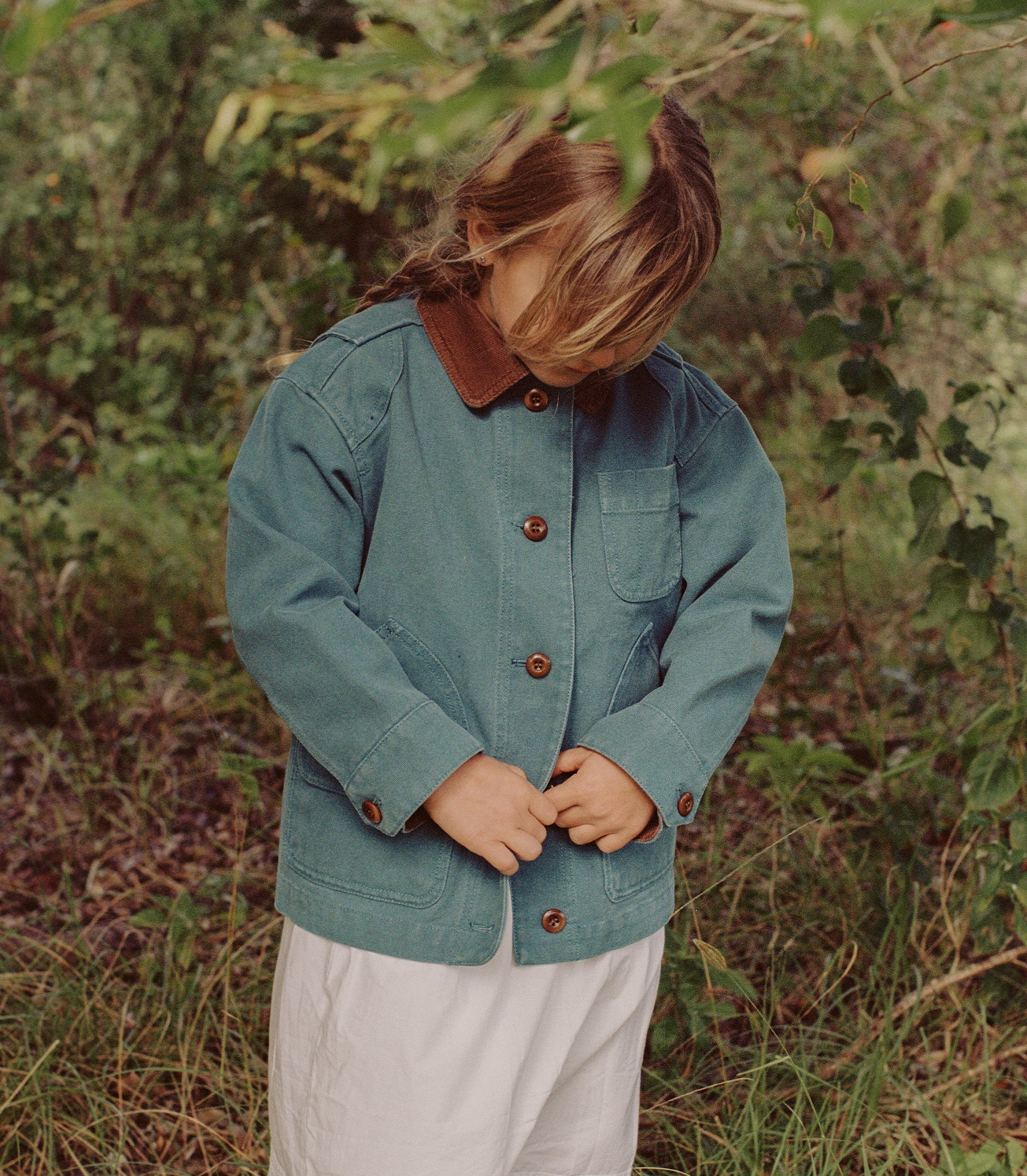A child wearing a teal blue denim jacket with a brown corduroy collar and brown buttons, standing outdoors in a grassy, wooded area.