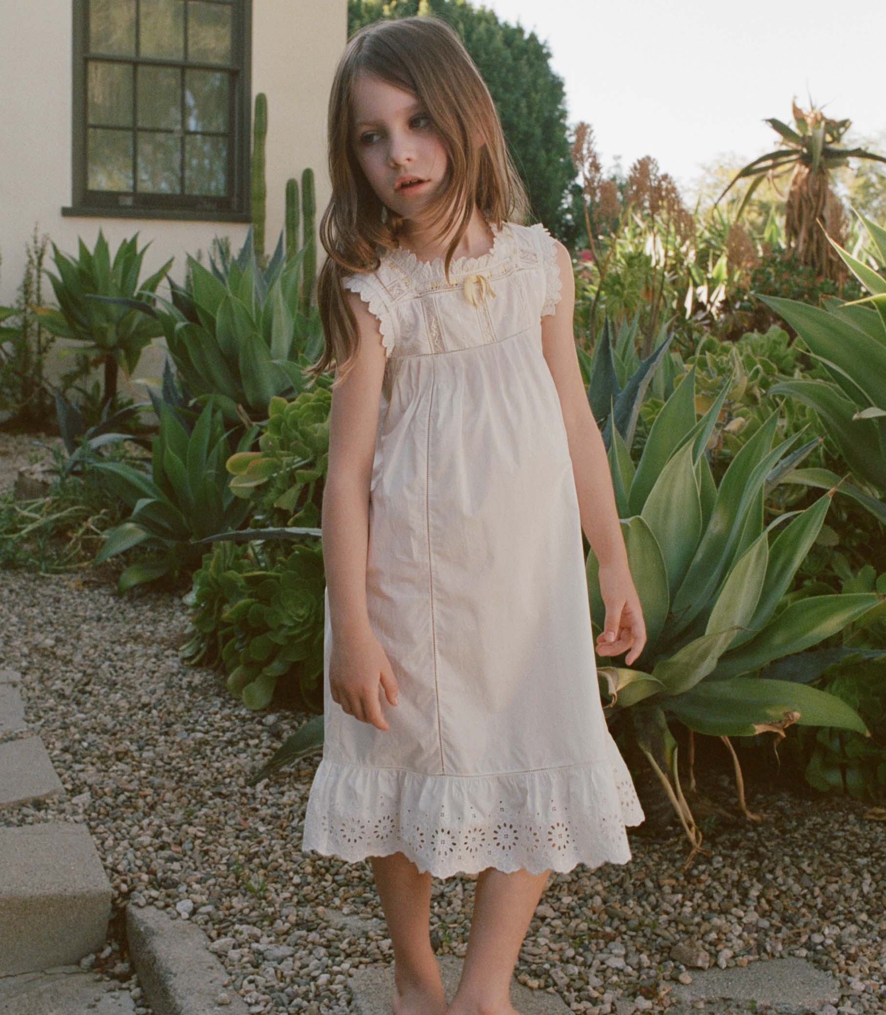 Young girl in a white dress standing in a garden with green plants and a building in the background. 1
