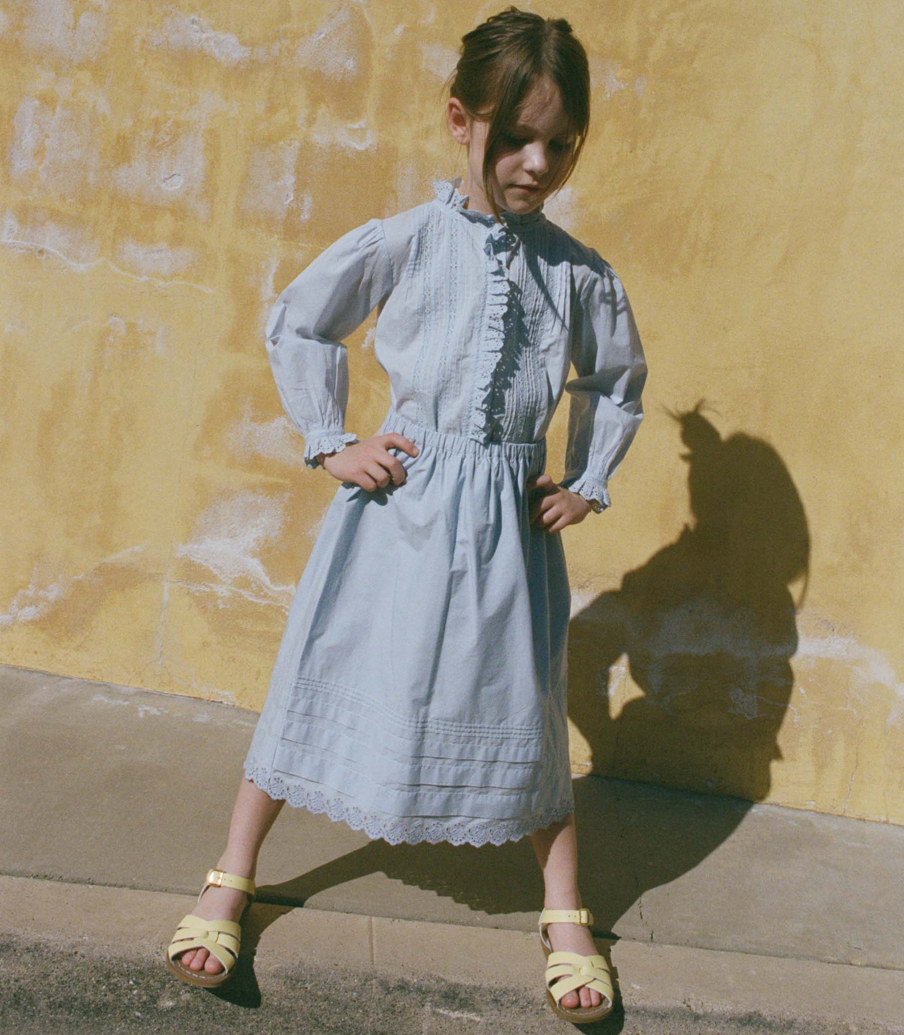 Little girl in a light blue skirt with a matching blouse standing against a yellow wall. 1
