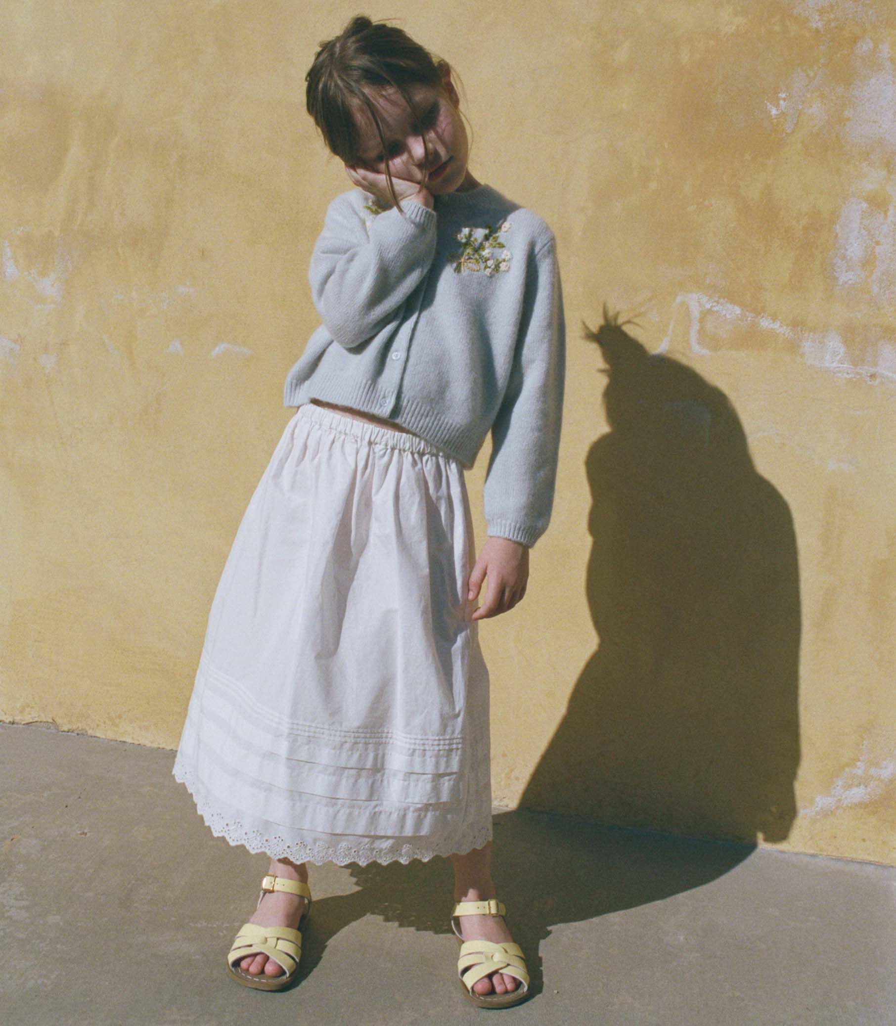 Little girl wearing a light blue sweater and white skirt against a yellow wall. 1
