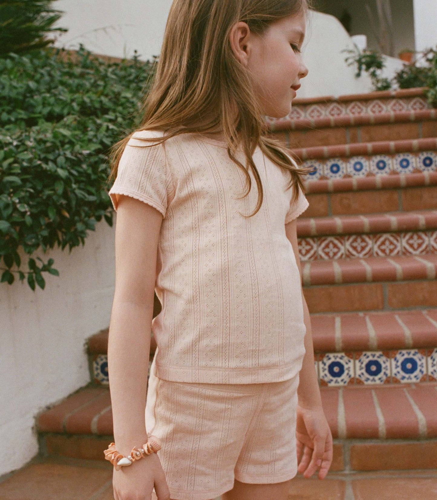 Young girl in a peach outfit standing on a tiled staircase with greenery in the background