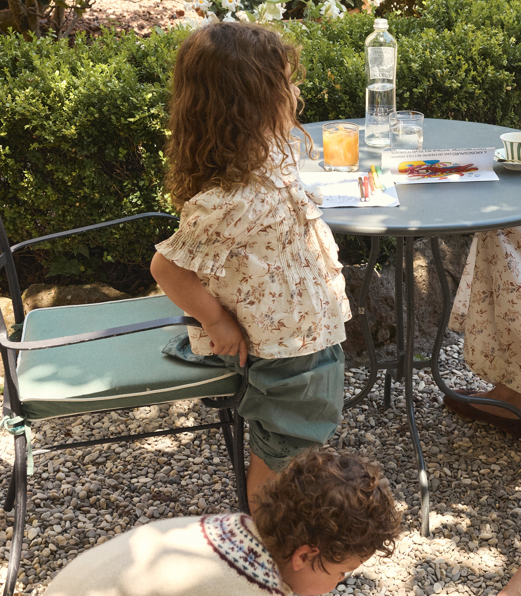 A child stands at an outdoor patio table wearing a cream floral ruffled blouse and green shorts, with another child kneeling in the foreground on a gravel surface.