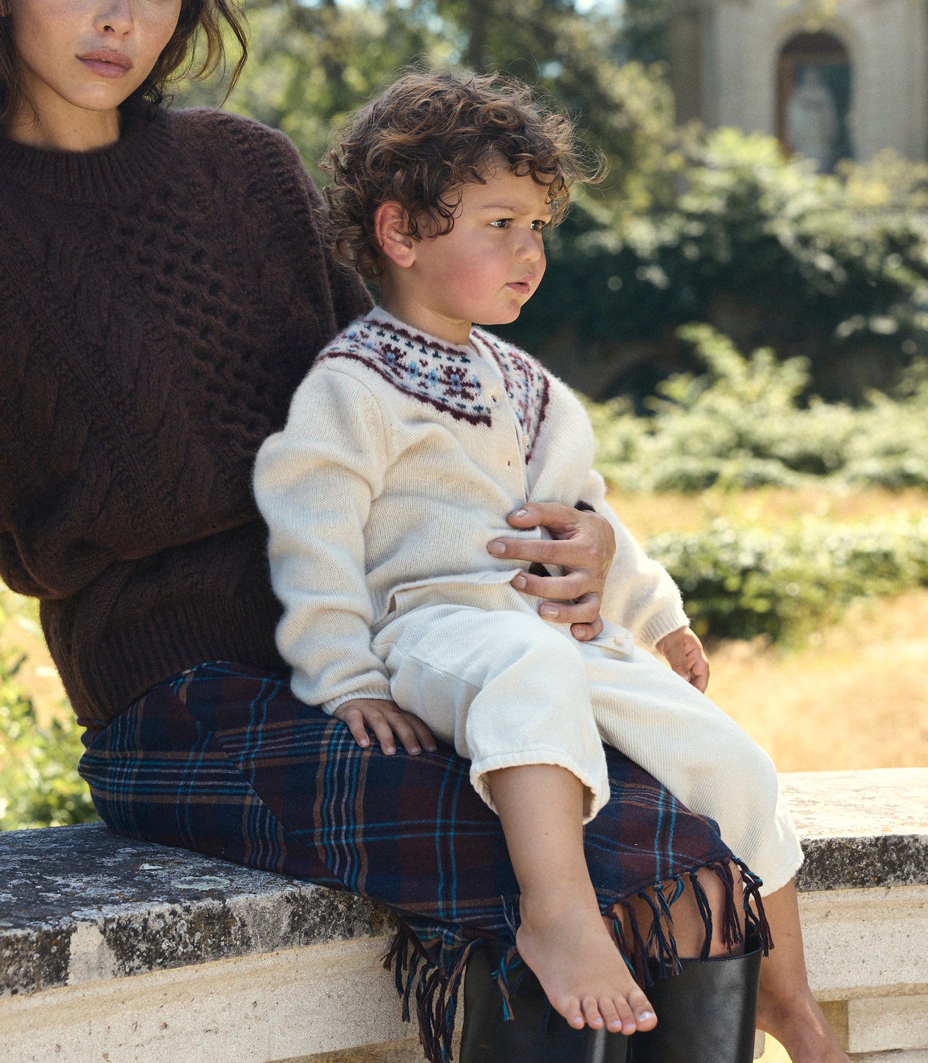 A child wearing a cream knit sweater and matching pants sits on an adult's lap outdoors. The child's outfit has a colorful embroidered yoke at the neckline.