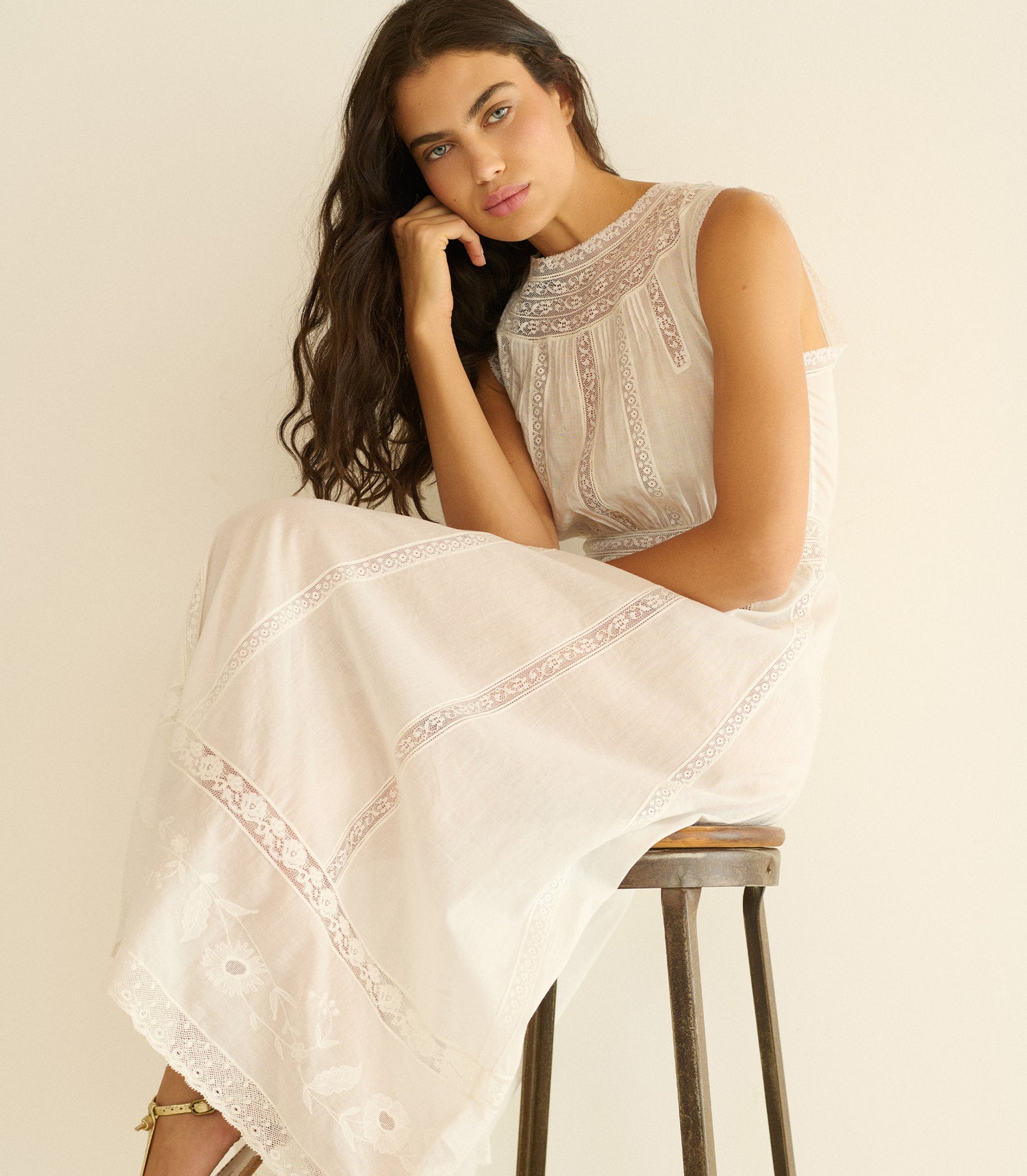 Woman sitting on a stool wearing a white lace dress against a plain background