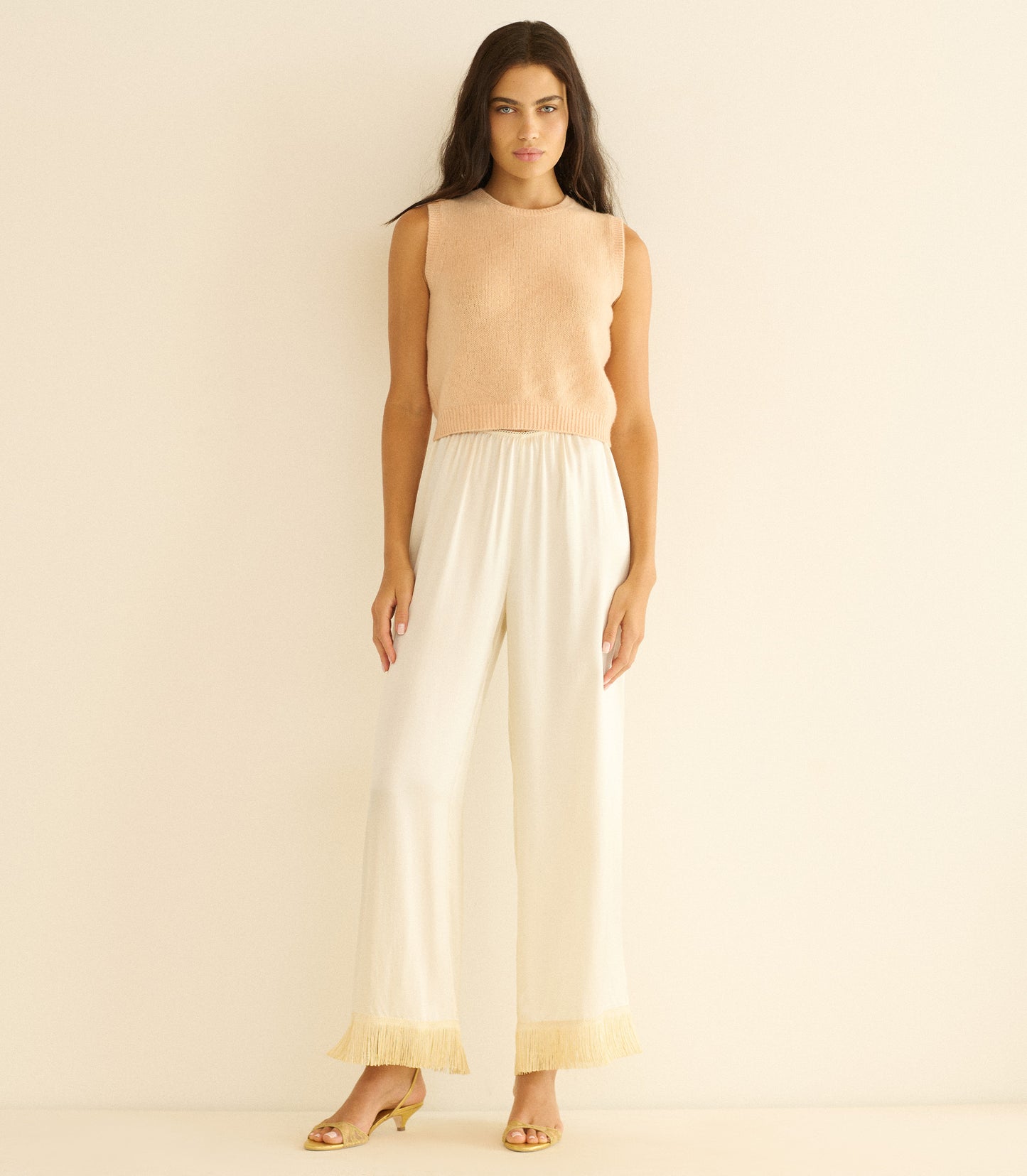 Woman wearing a peach sleeveless top and white pants against a beige background