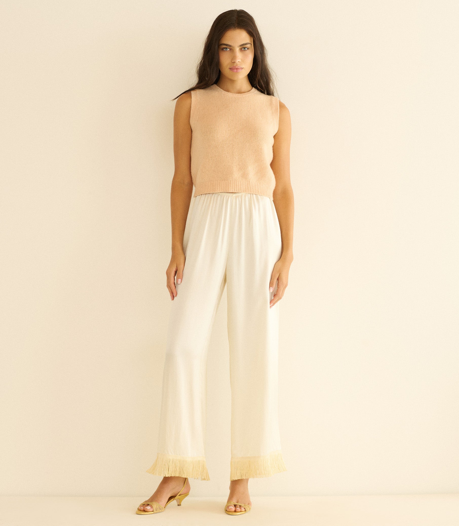 Woman wearing a peach sleeveless top and white pants against a beige background 2
