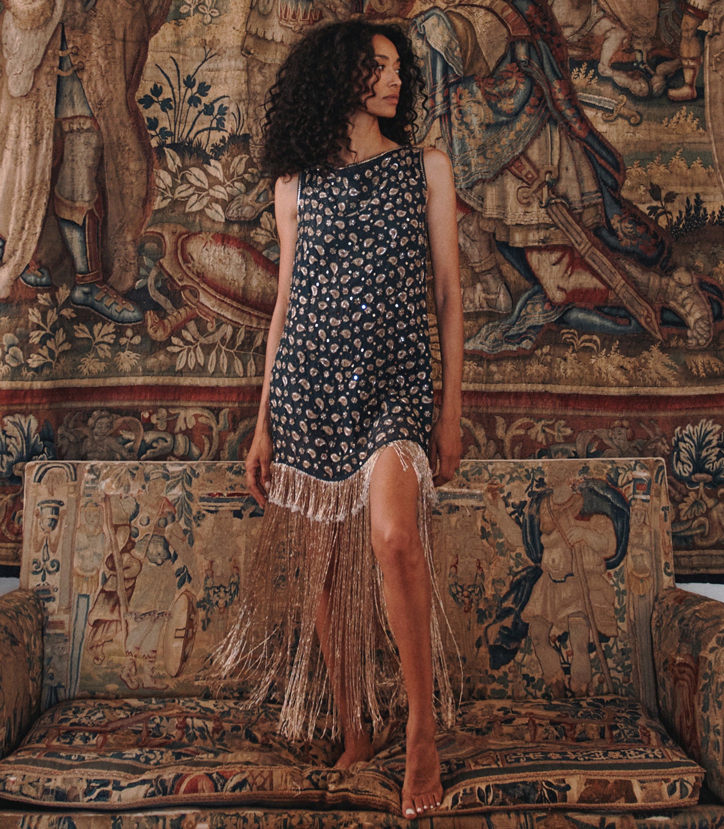 A woman stands barefoot on an ornate tapestry sofa wearing a sleeveless dark dress with a small floral print and long fringe along the hem, posed in front of a large matching tapestry wall hanging.