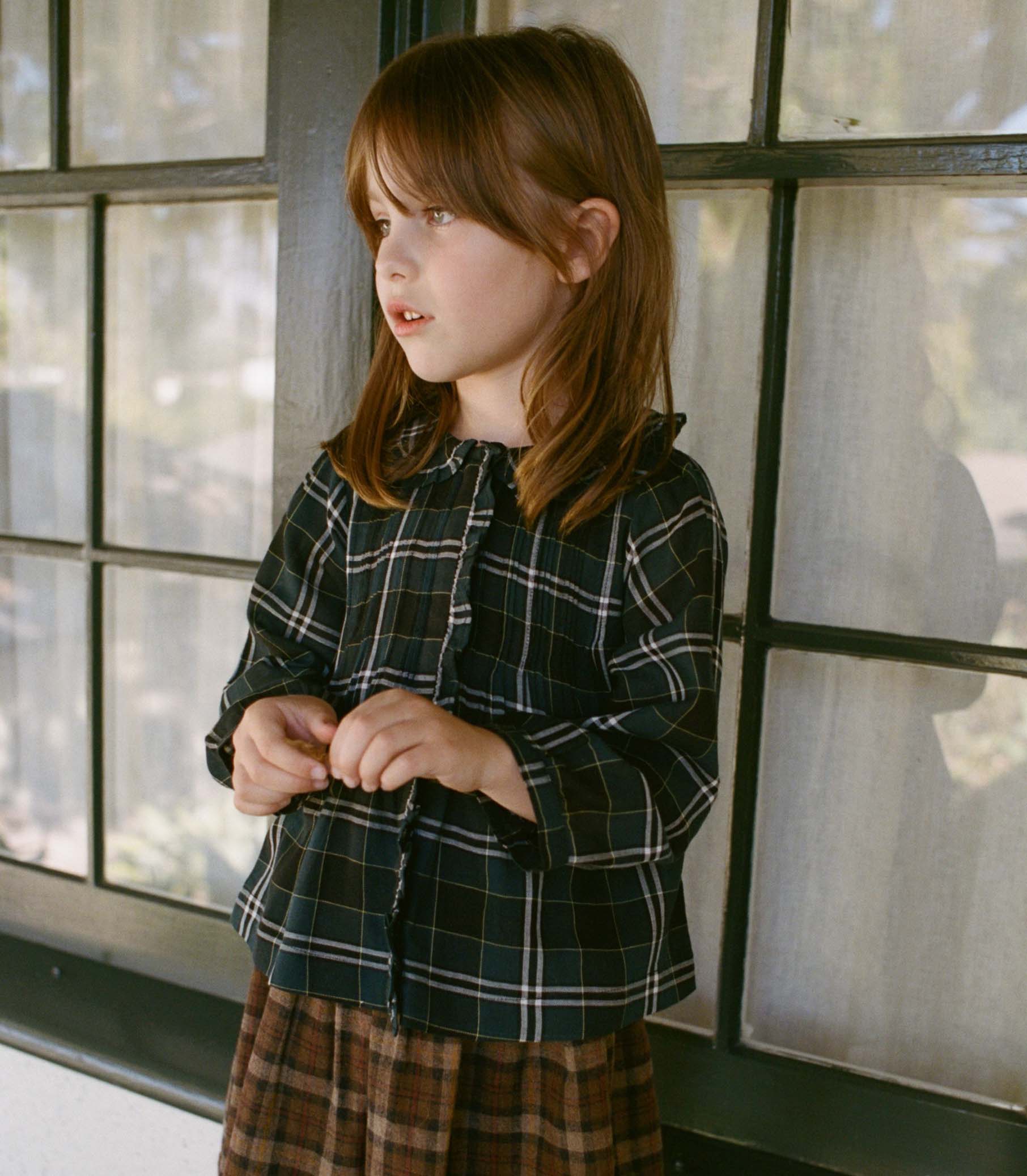 Young girl wearing a plaid shirt and skirt standing in front of a window. 1
