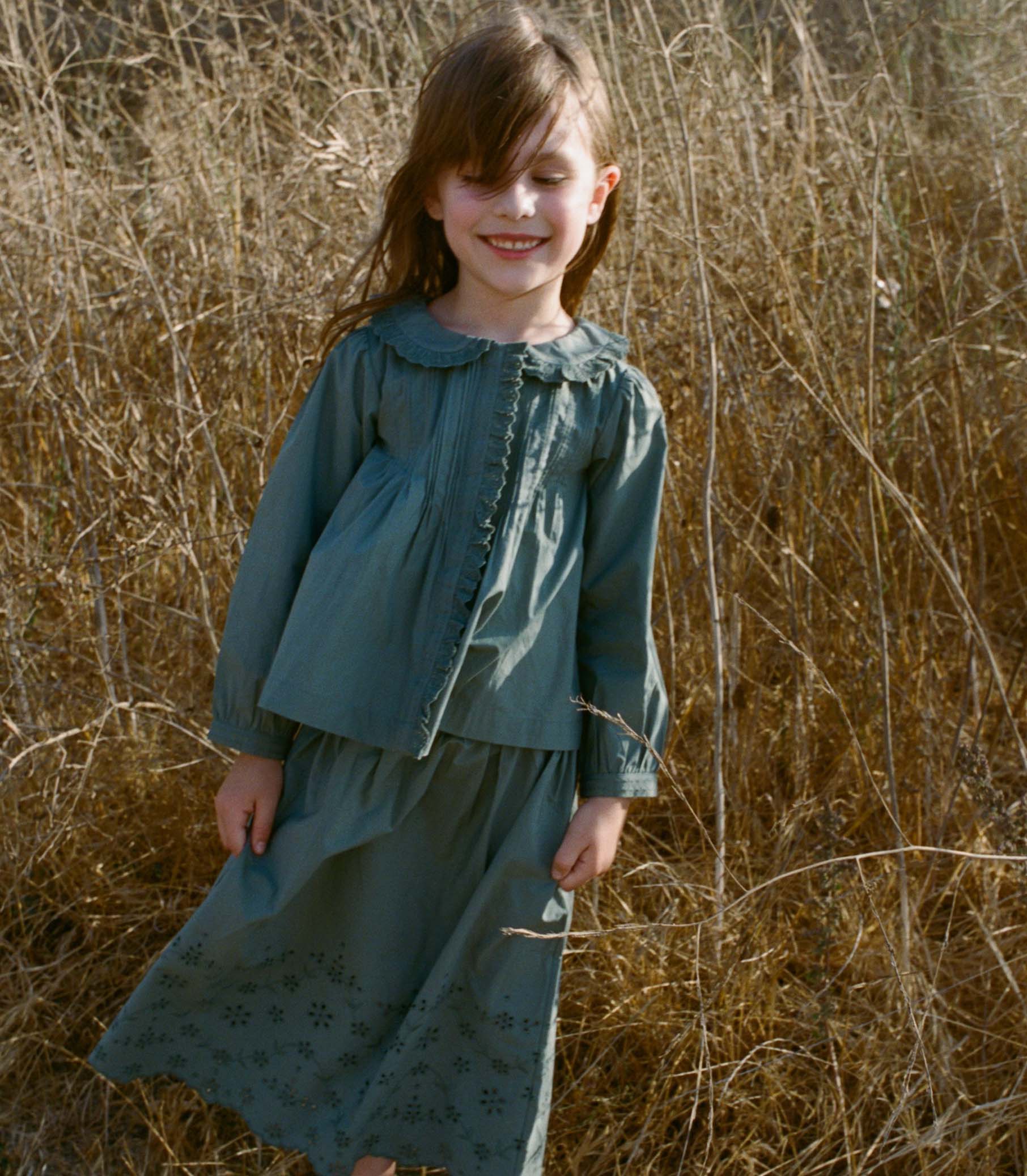 Young girl in a teal top and matching skirt, standing in a field of dry grass. 1
