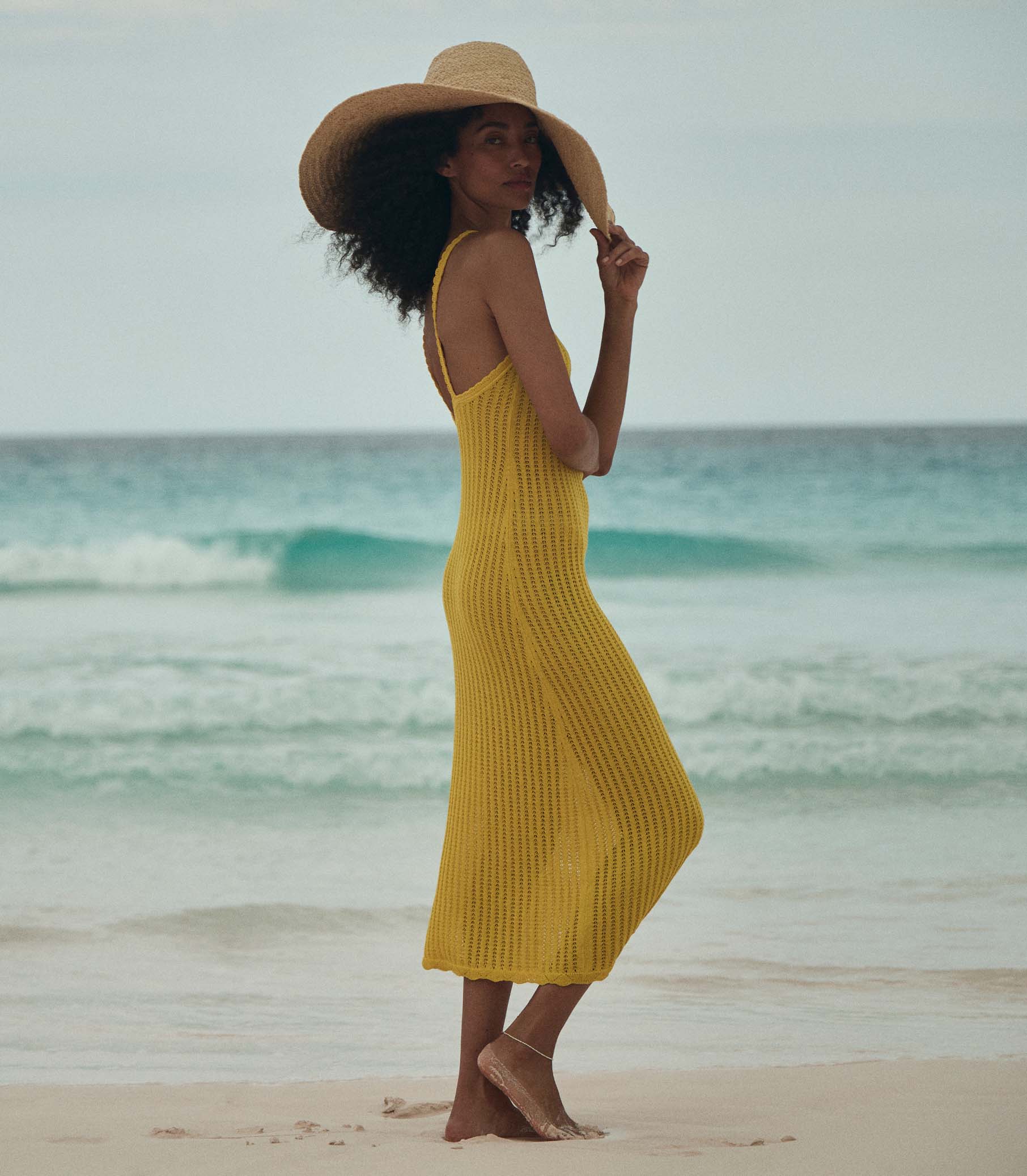 Anais is wearing a sleeveless yellow knit maxi dress with a sun hat on the beach. 3
