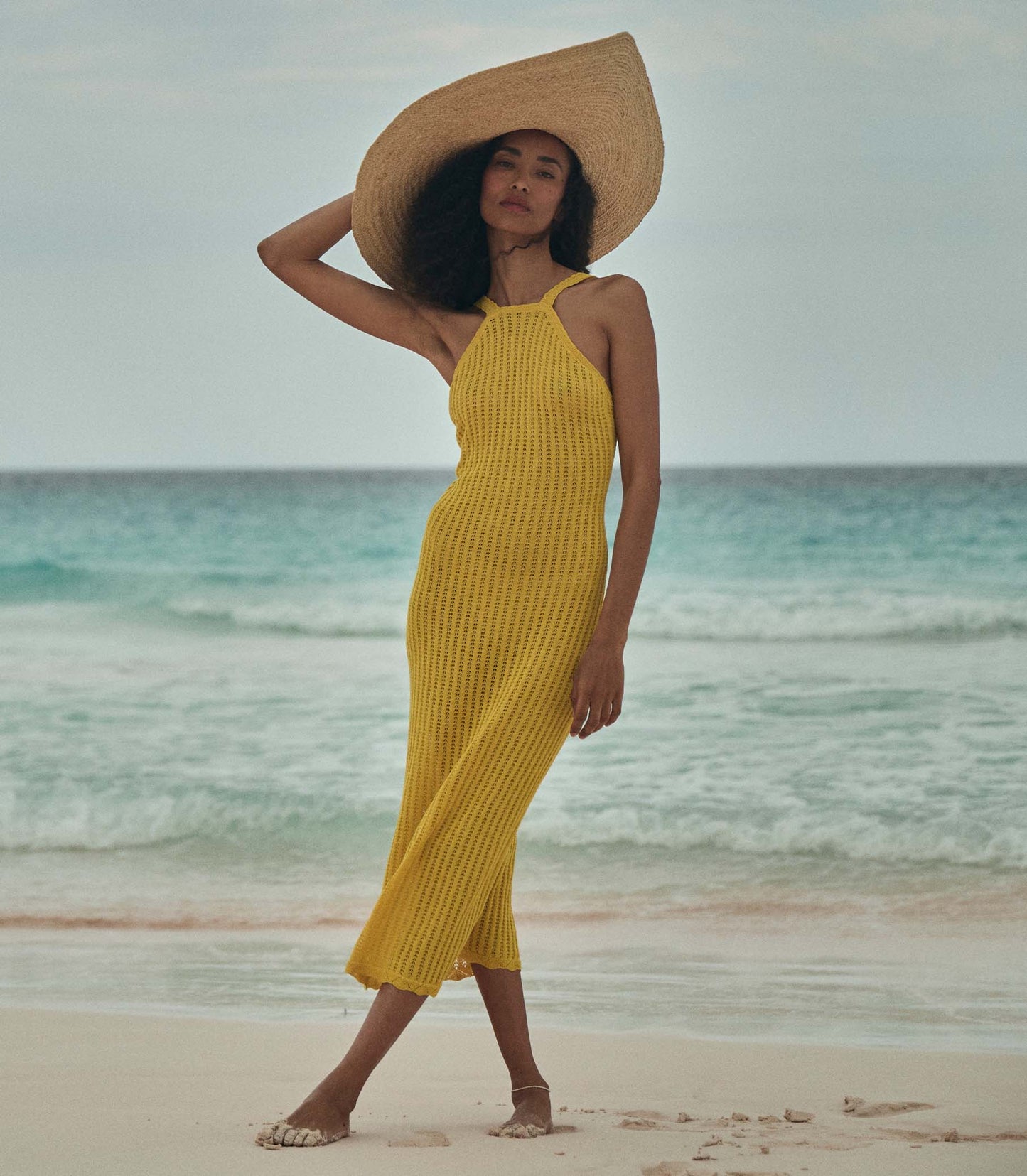 Anais is wearing a sleeveless yellow knit maxi dress with a sun hat on the beach.